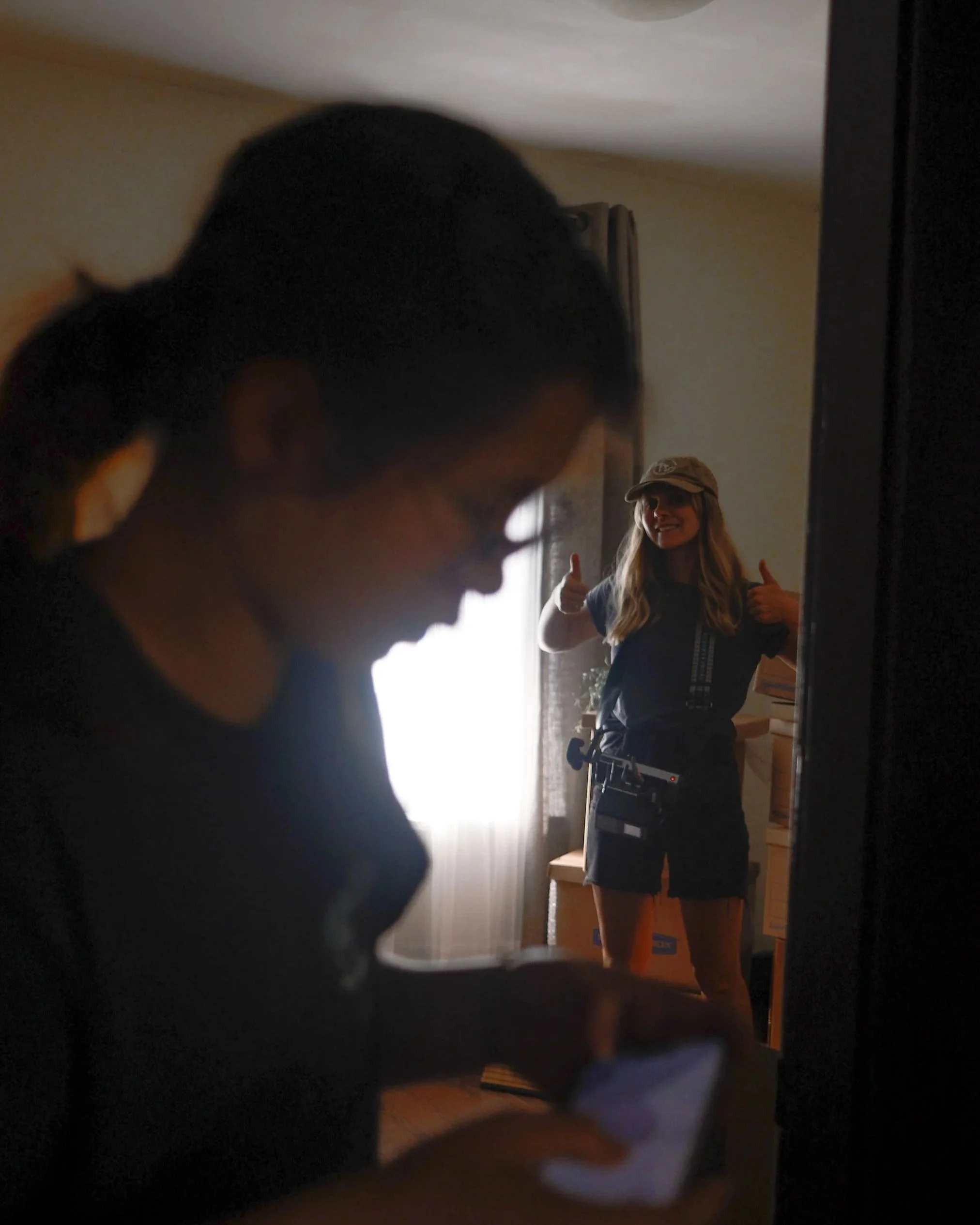 Two women inside a room; one woman is in the foreground, looking at her phone, blurred; the other woman is in the background, smiling and giving two thumbs up, wearing a hat and a harness.