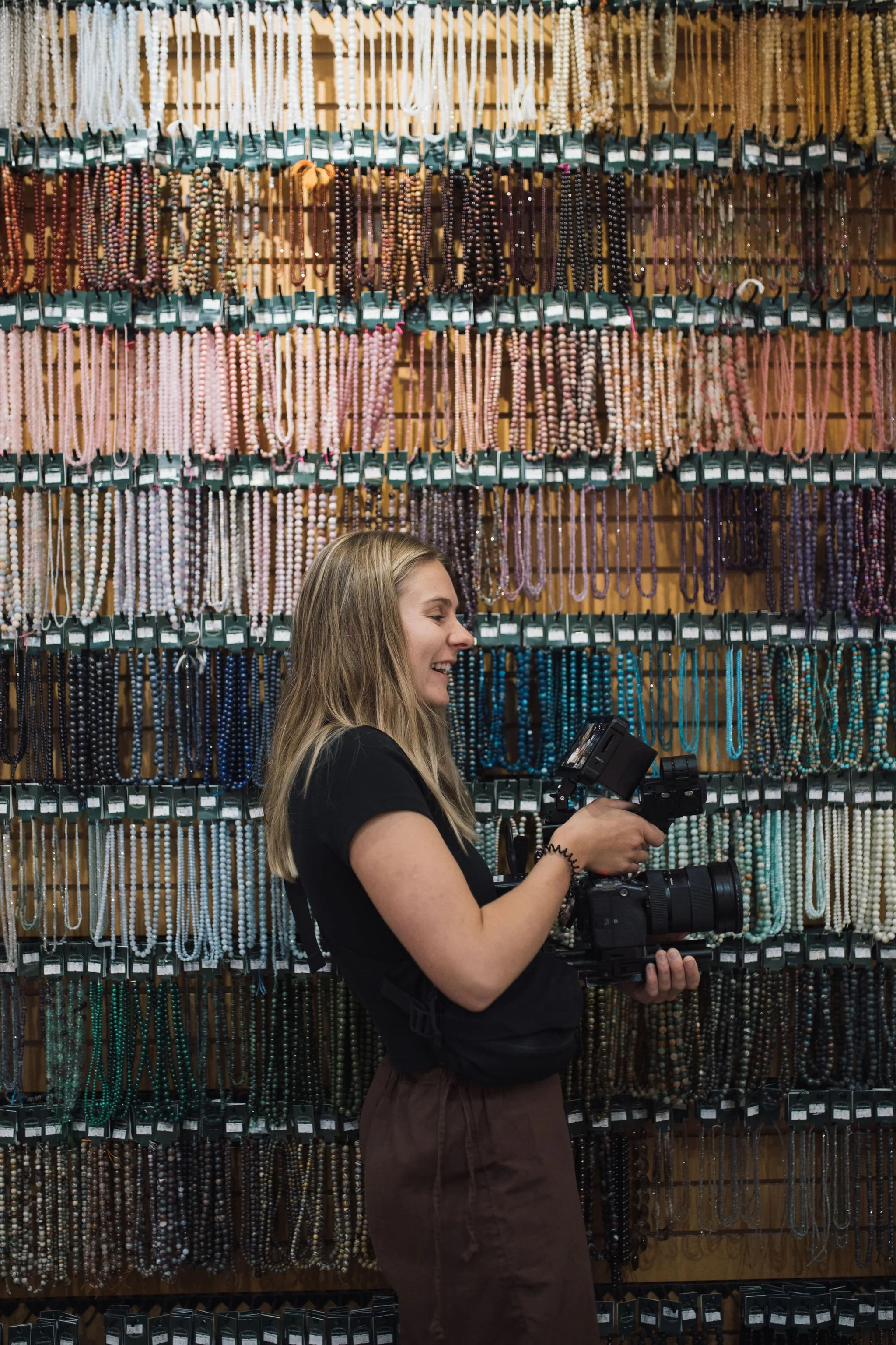 A woman shopping for necklaces at a store. She is smiling and holding a camera, standing in front of a wall filled with colorful beaded necklaces.