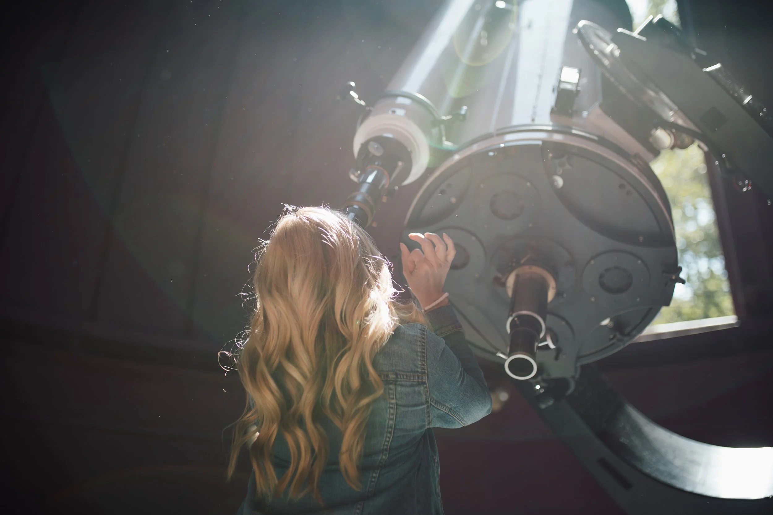 A woman with long blonde hair, wearing a denim jacket, is looking at and reaching towards the eyepiece of a telescope in a dark room with sunlight coming through a window.