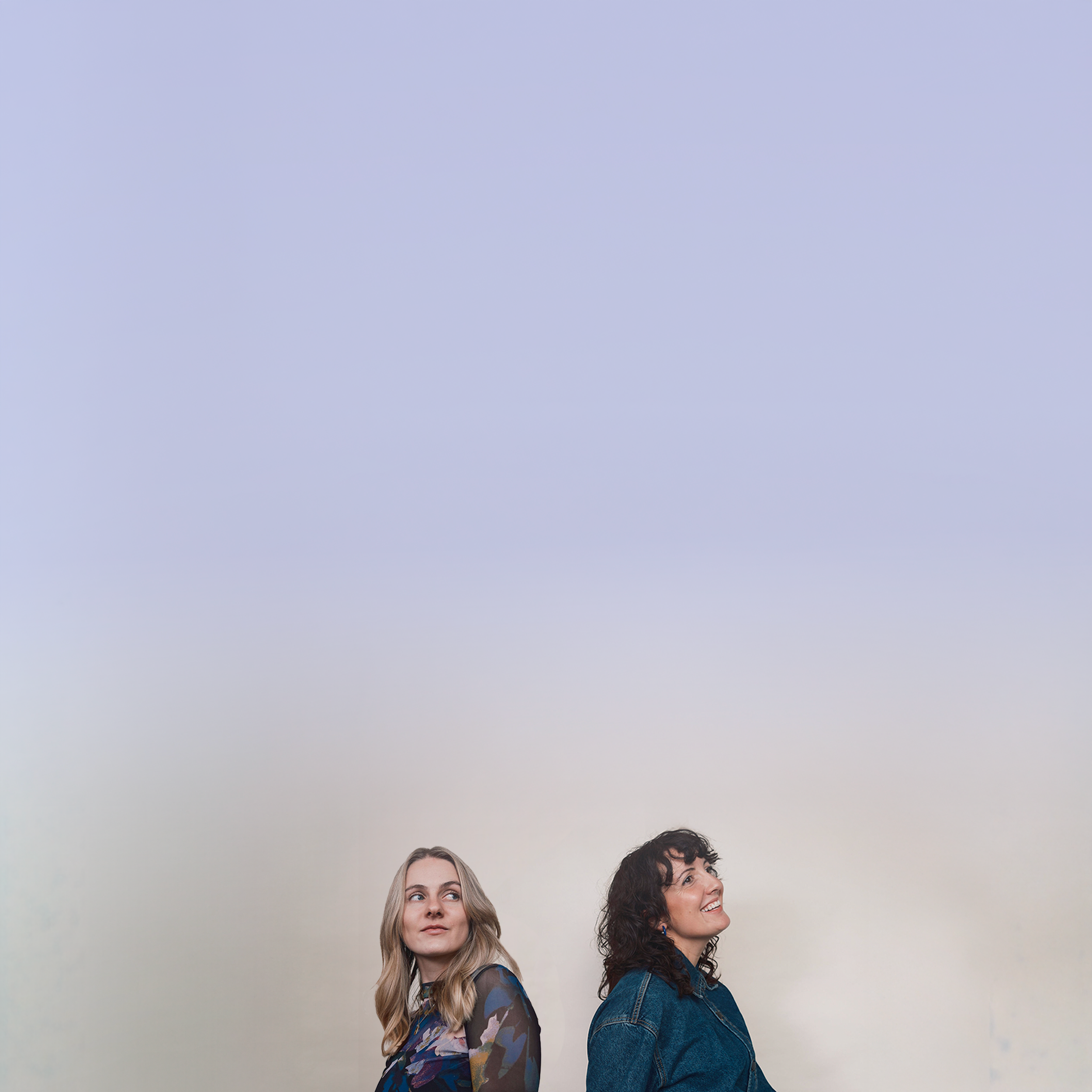 Two women standing back-to-back, looking in opposite directions, against a plain wall.