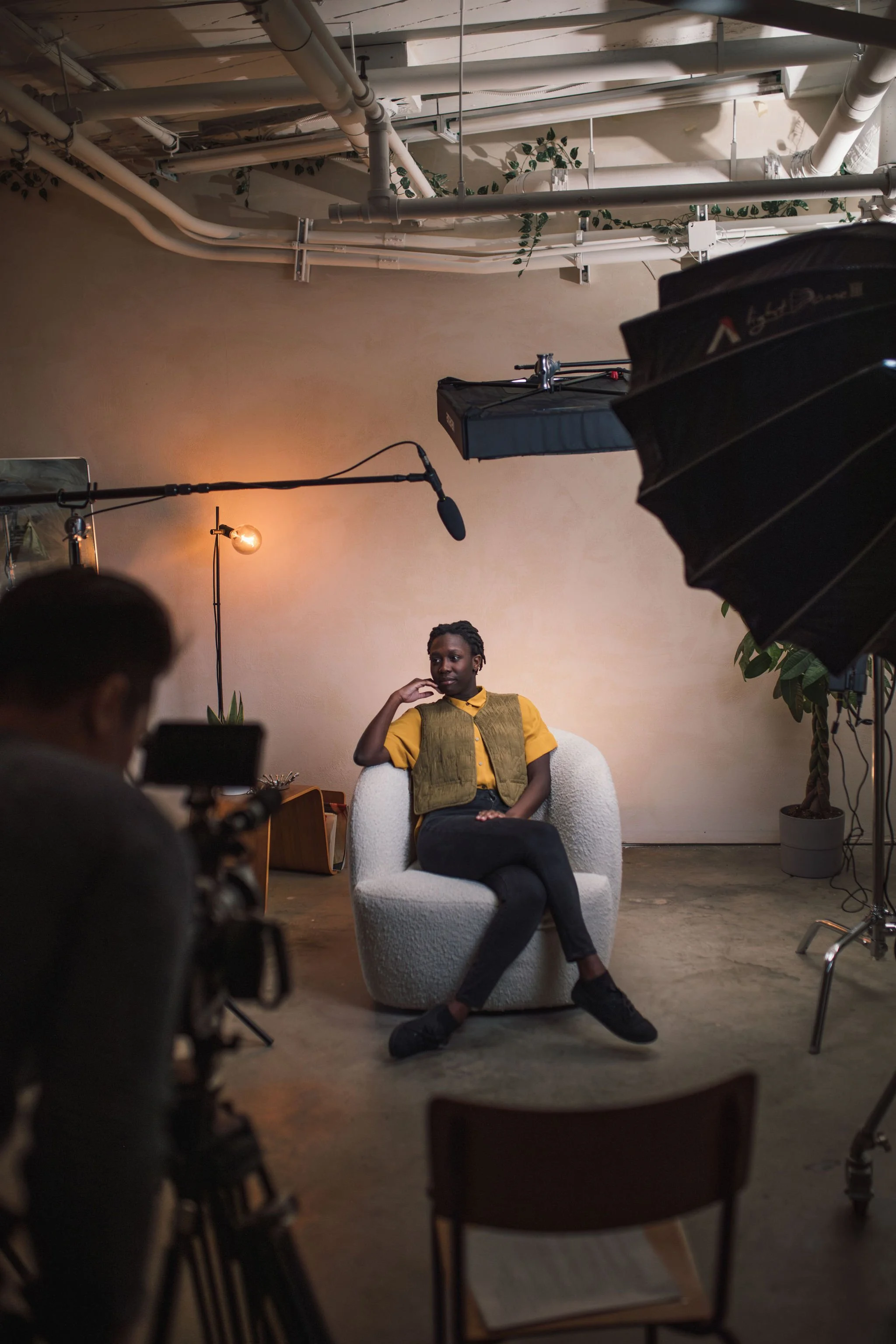 A woman sitting on a plush white armchair during a video shoot or interview, with film equipment and lighting around her in a studio with minimal decor and potted plants.