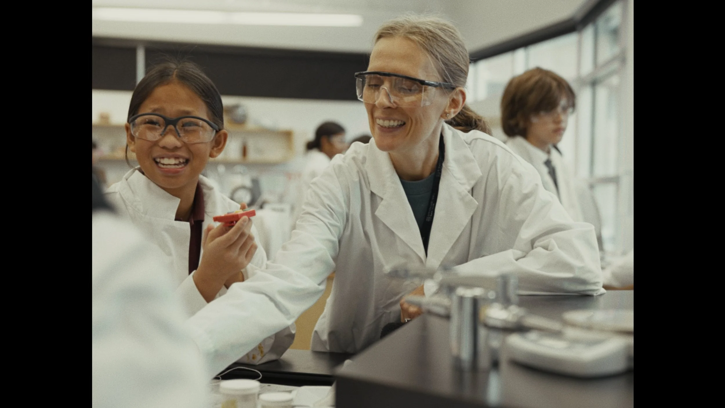 A teacher and a student in a science classroom, both wearing safety goggles and lab coats, smiling and working together.