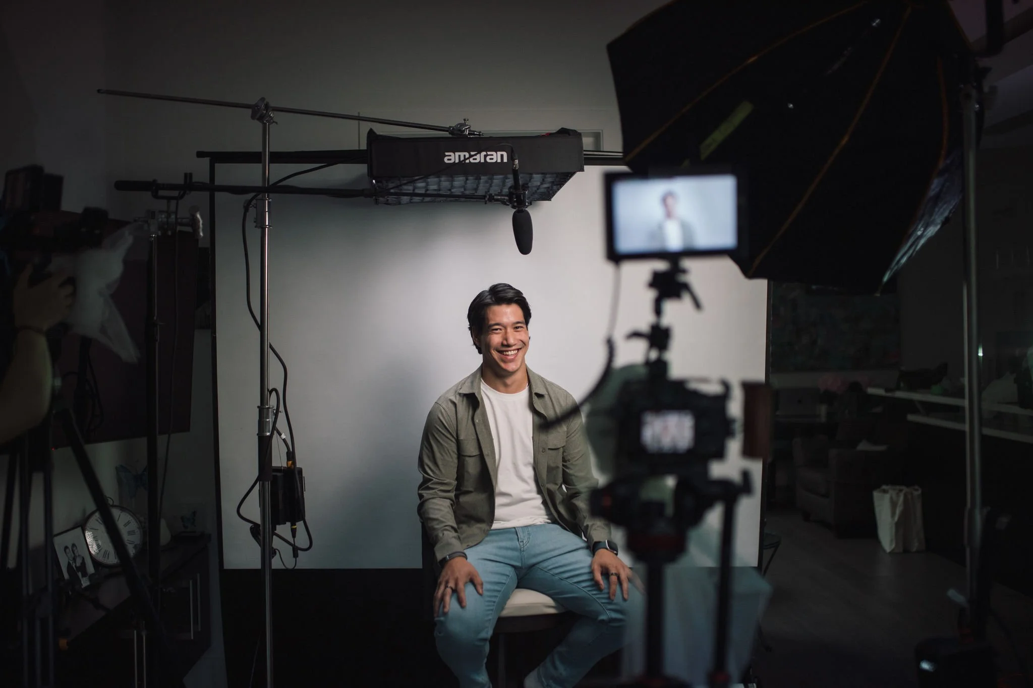 Young man sitting on a chair in a video recording studio, smiling for the camera, with professional lighting and camera equipment around him.