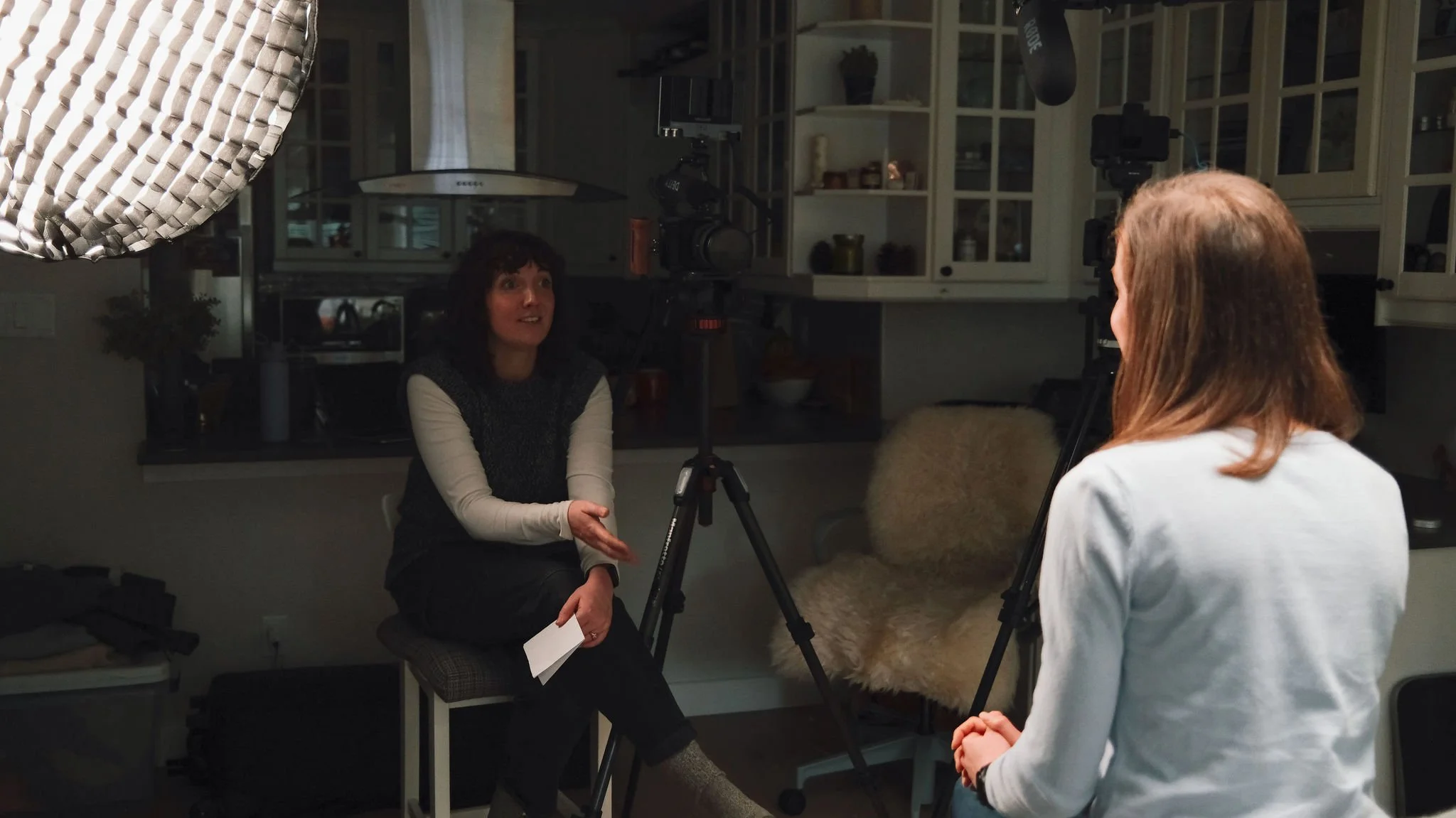 Two women conducting an interview or recording a conversation in a home kitchen. One woman sits on a chair with a notepad, the other stands facing her. Camera equipment and lighting are set up for recording.
