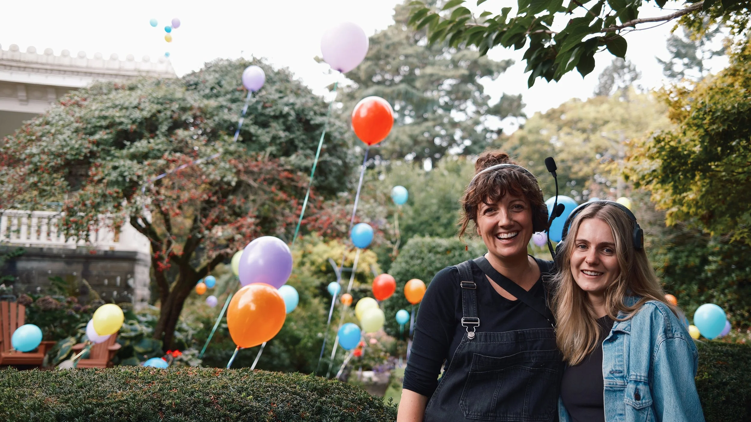 Two women smiling, standing outdoors in a garden with colorful balloons floating in the background, surrounded by trees and greenery.