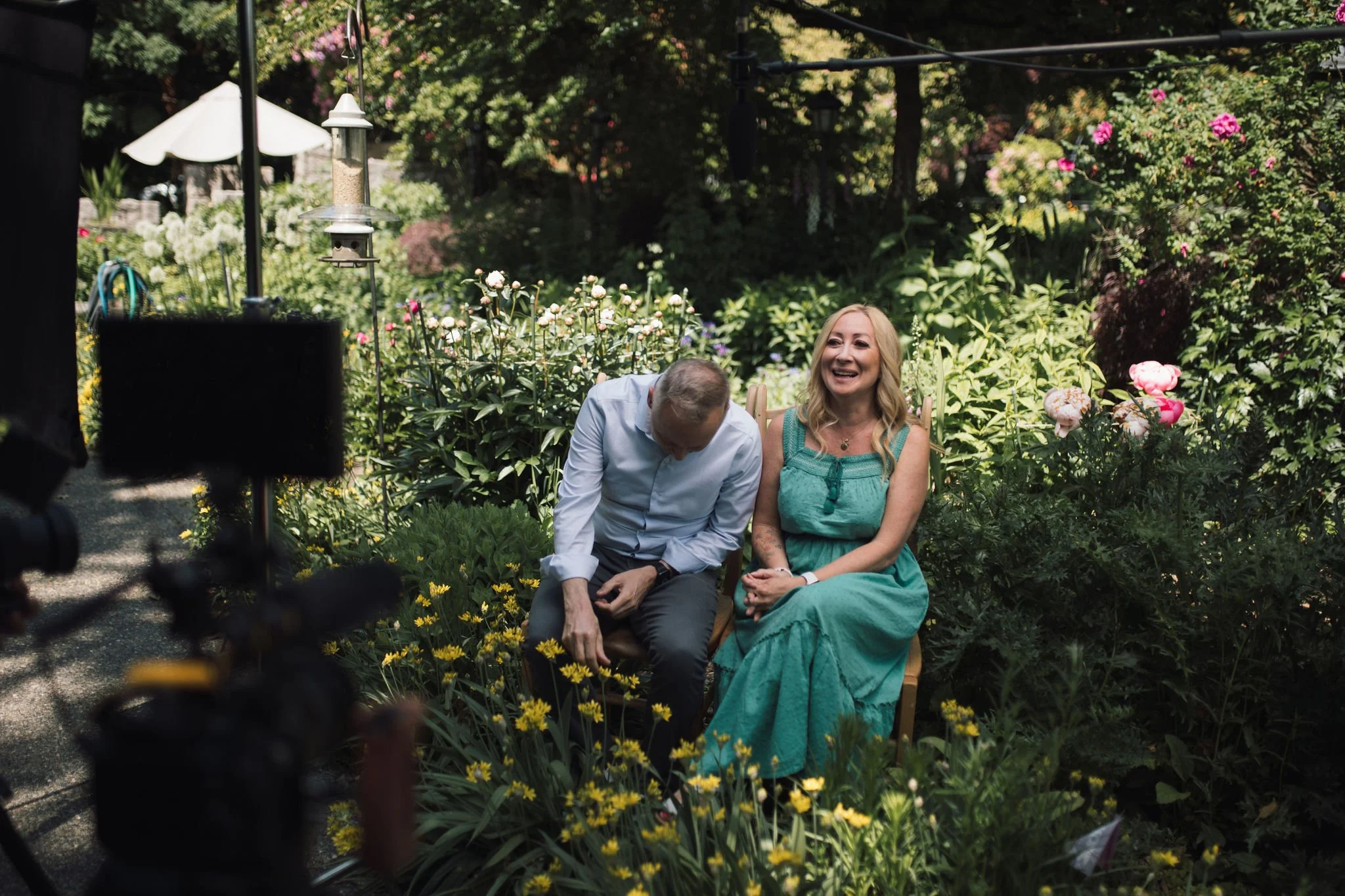 A man and a woman sit on a bench in a lush garden, laughing and smiling, with filming equipment visible in the foreground.