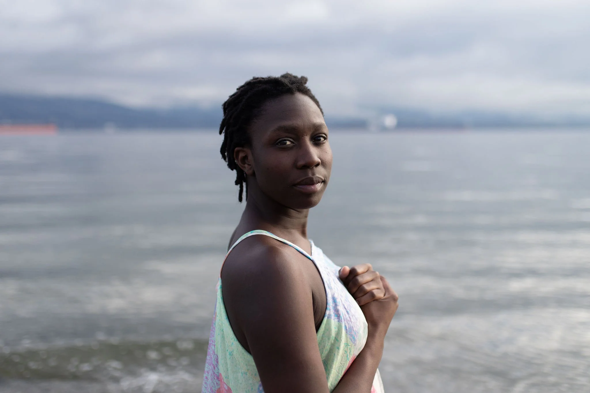 A woman with short dreadlocks stands by the water on a cloudy day, wearing a pastel-colored dress, looking at the camera with a calm expression.