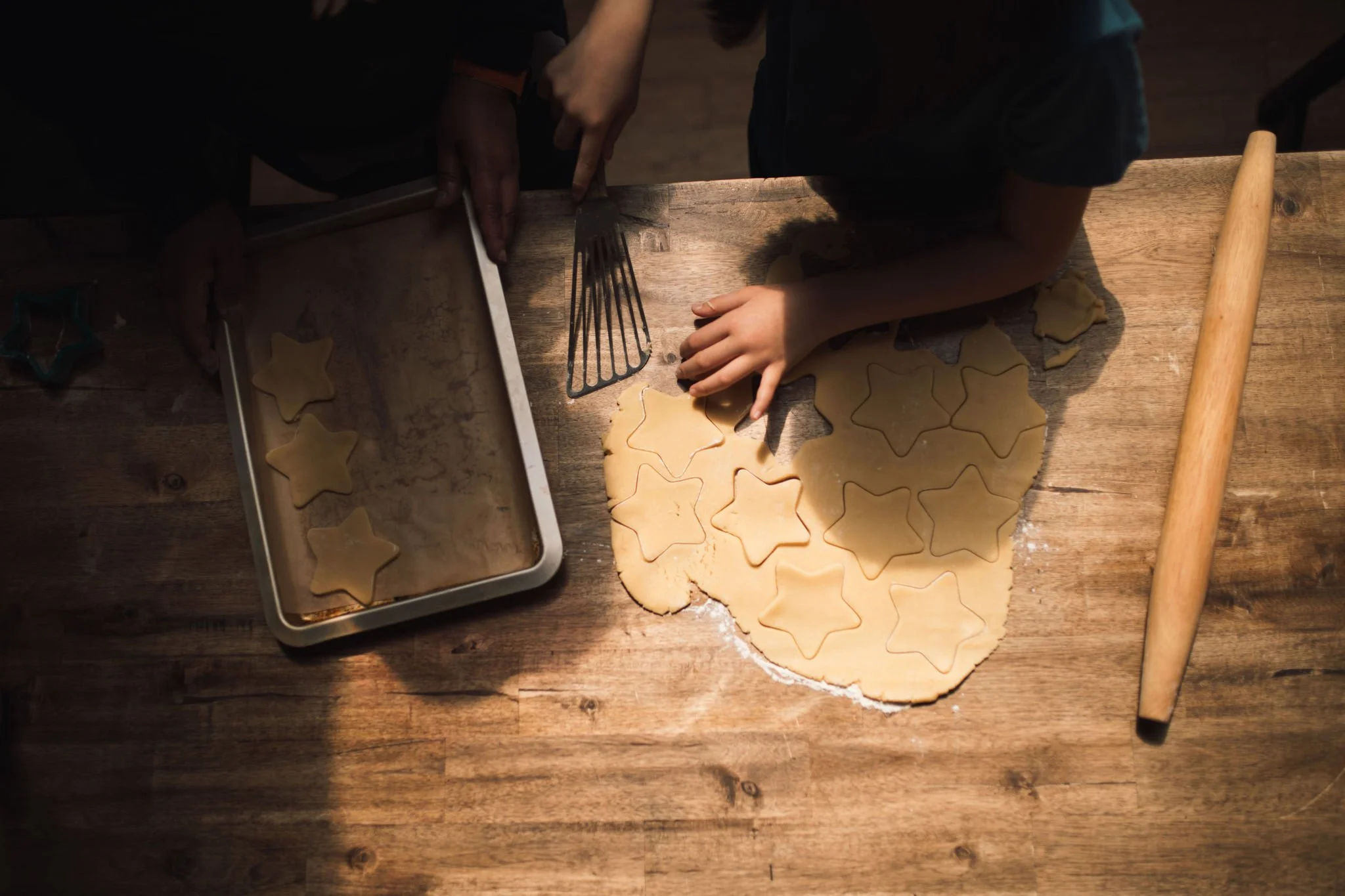 Two children are making star-shaped cookies. One child is using a cookie cutter to cut out cookies from rolled dough, while the other is pointing at a cookie. There is a baking sheet with cookies on it and a rolling pin on the wooden table.