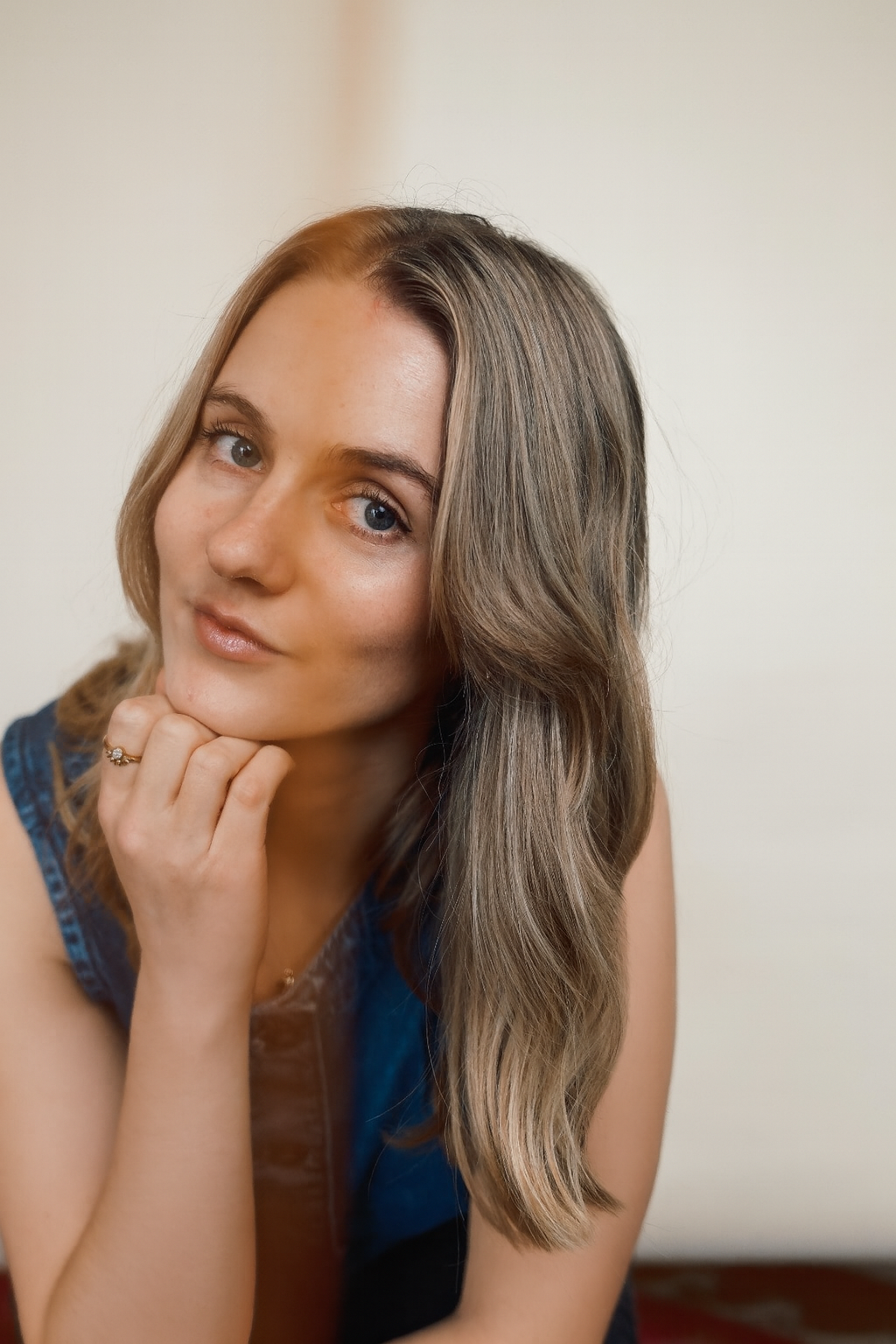 Close-up of a young woman with wavy brown hair, blue eyes, and a slight smile, wearing a blue denim top, resting her chin on her hand.