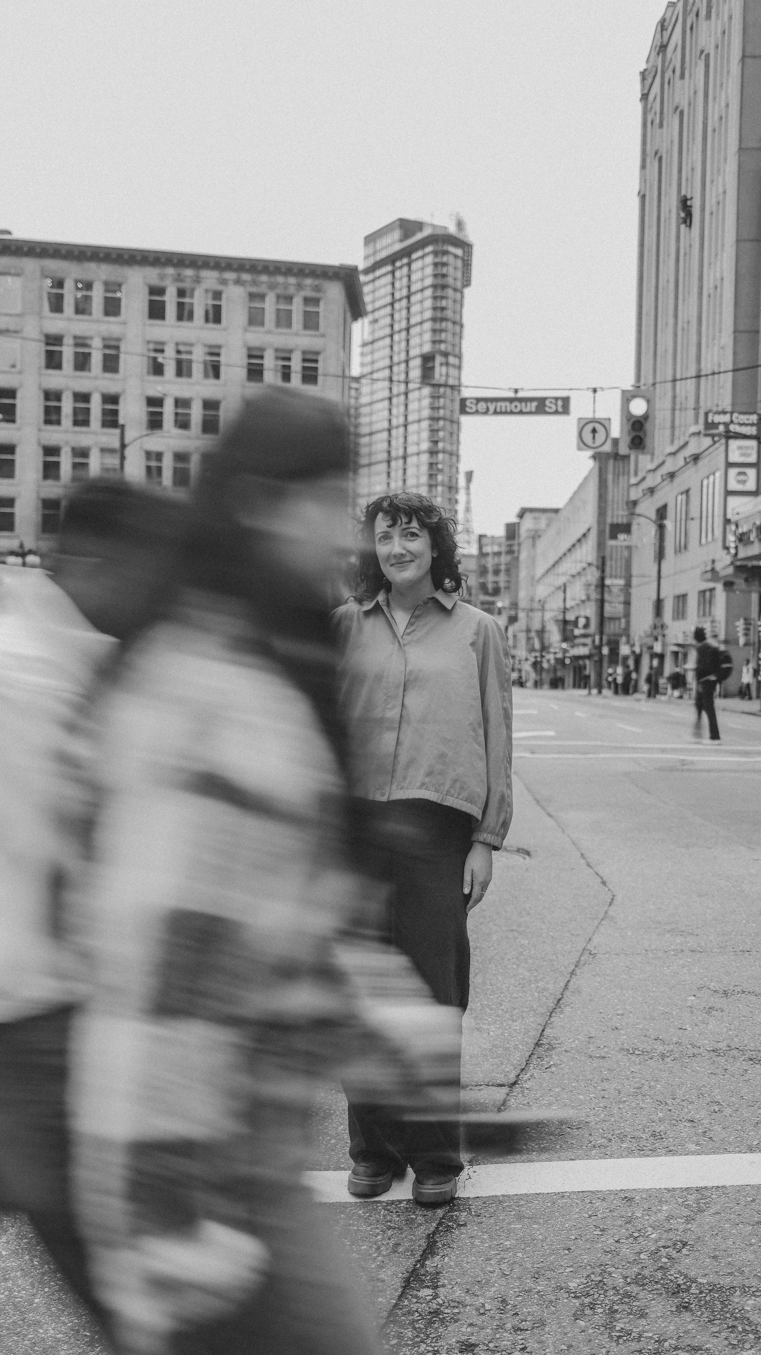 Black and white photo of a woman standing on a city street corner, with pedestrians and tall buildings in the background, under a street sign that says 'Seymour St'.