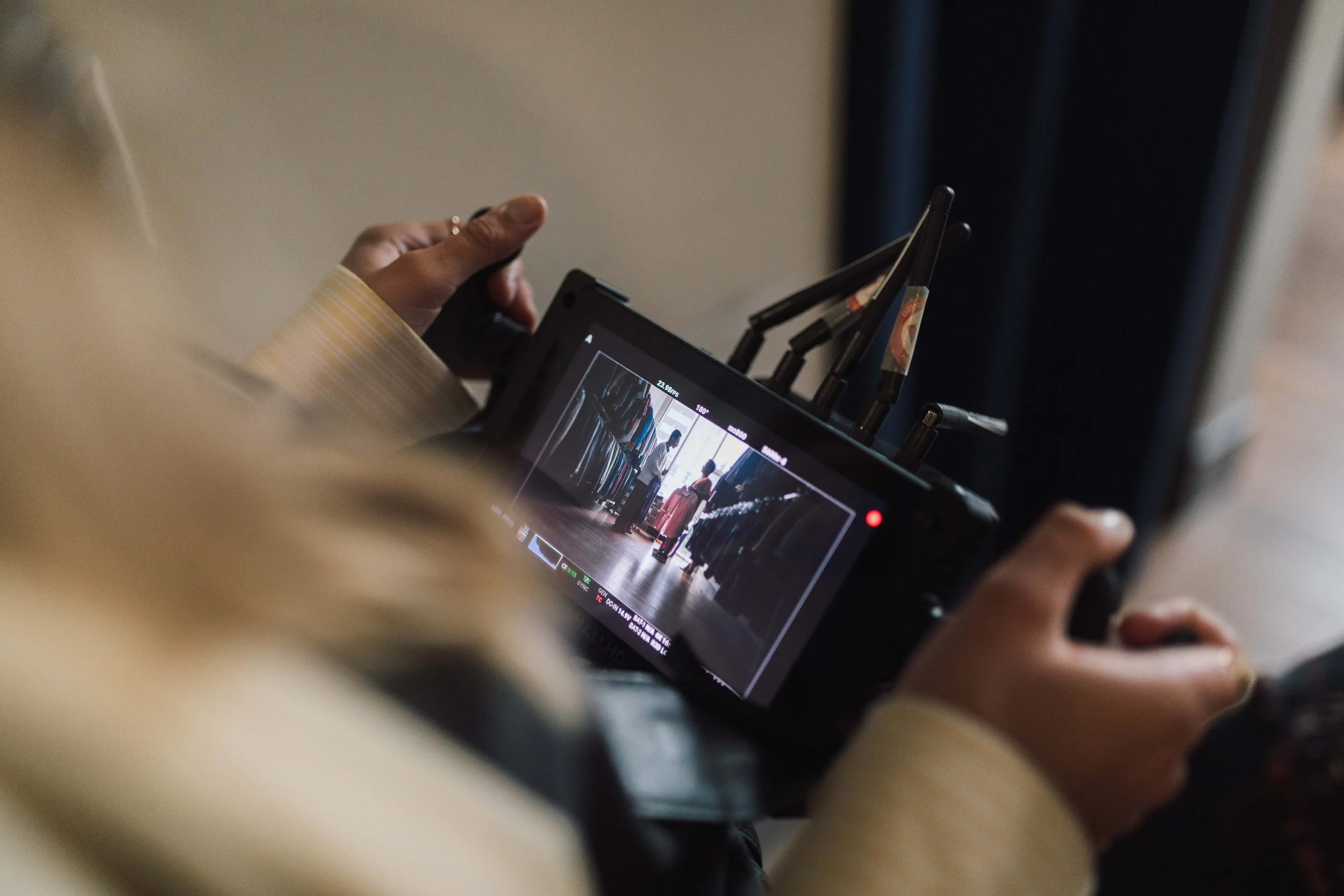 A person holding a film camera or monitor, capturing a scene of people at an airport terminal with luggage, seen on the screen of the camera.