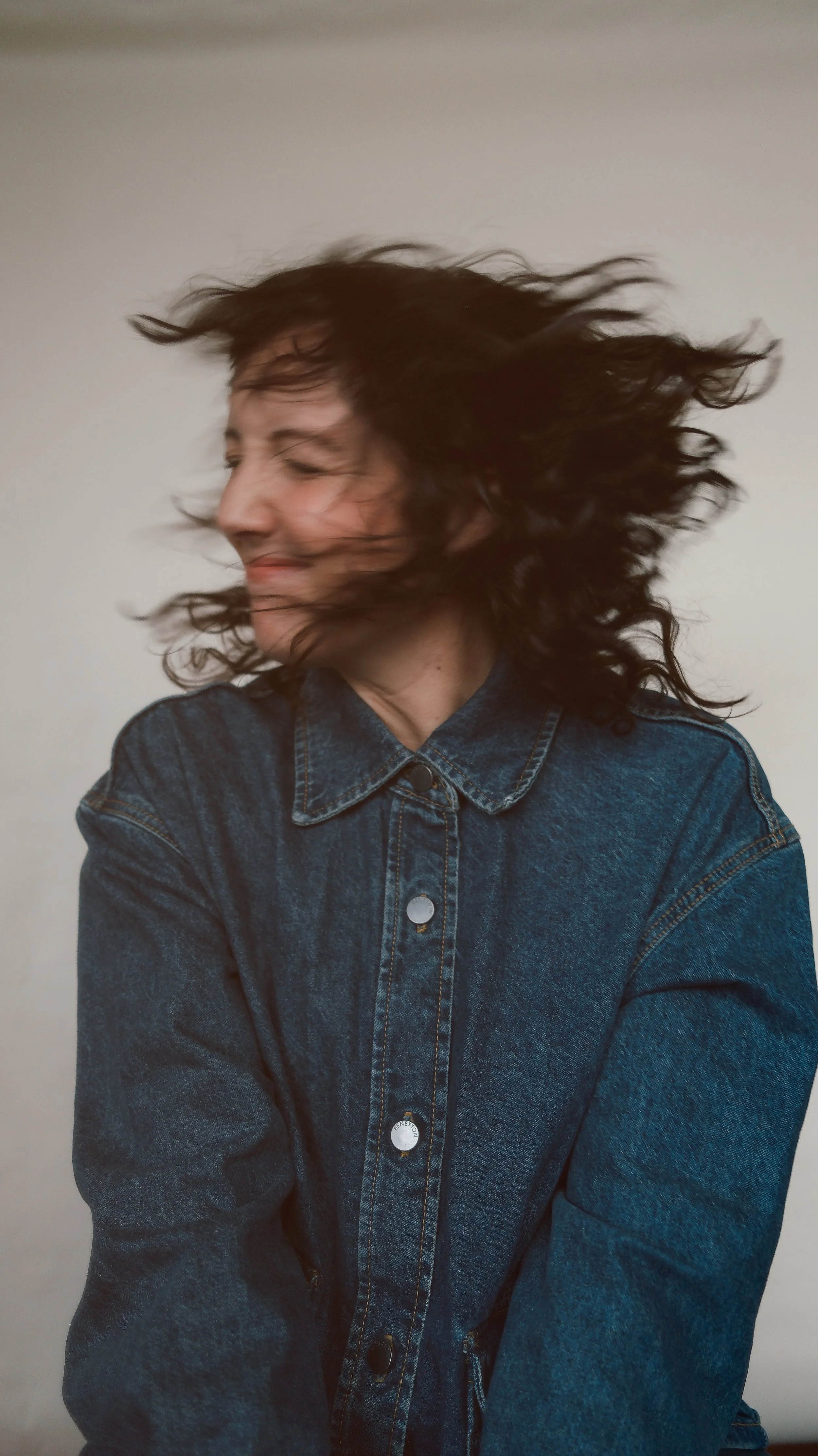 A woman with curly dark hair wearing a denim jacket, smiling with her eyes closed and hair blowing to the side.