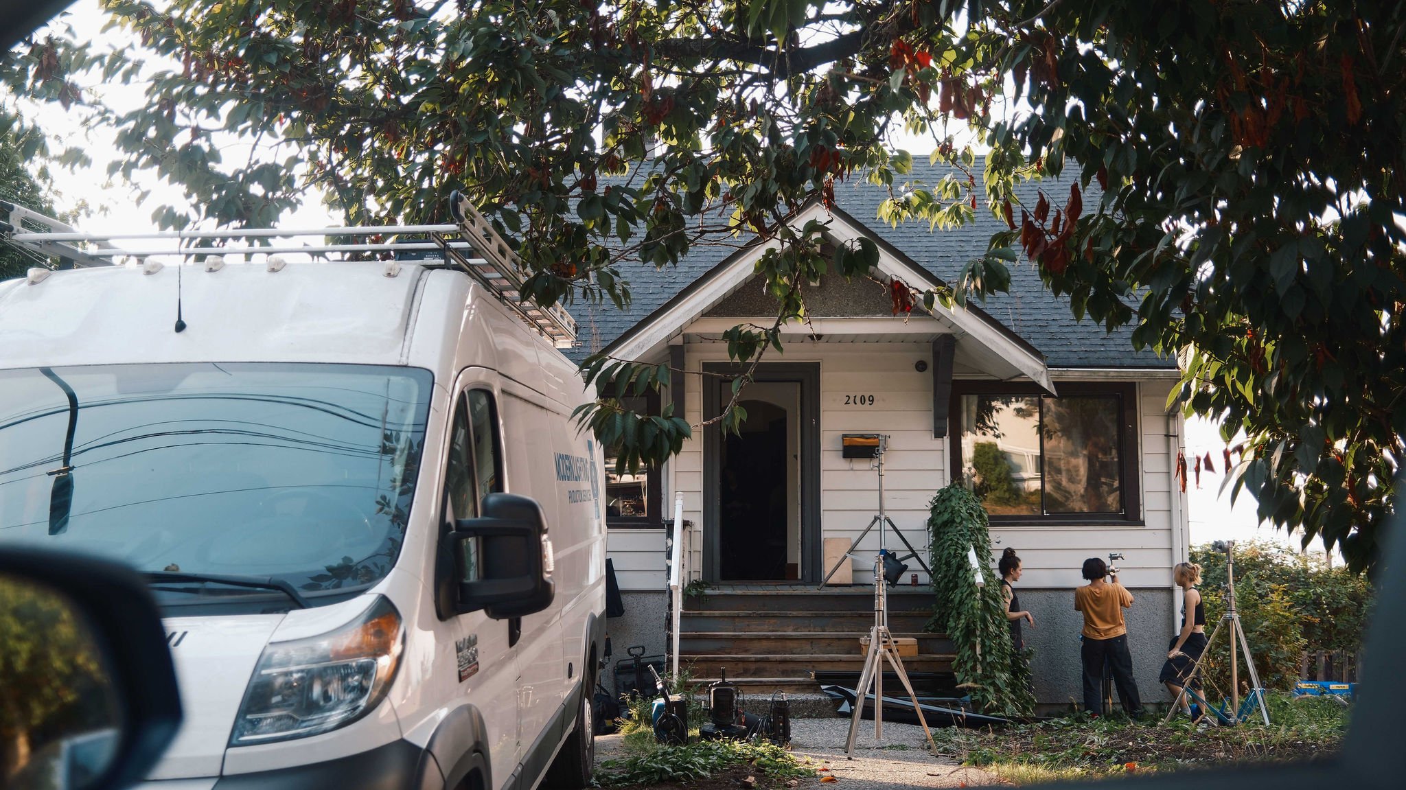 Scene of film or photo shoot setup in front of a house with three people engaged in conversation, surrounded by equipment including tripods and lighting, with a white van parked nearby and leafy trees overhead.