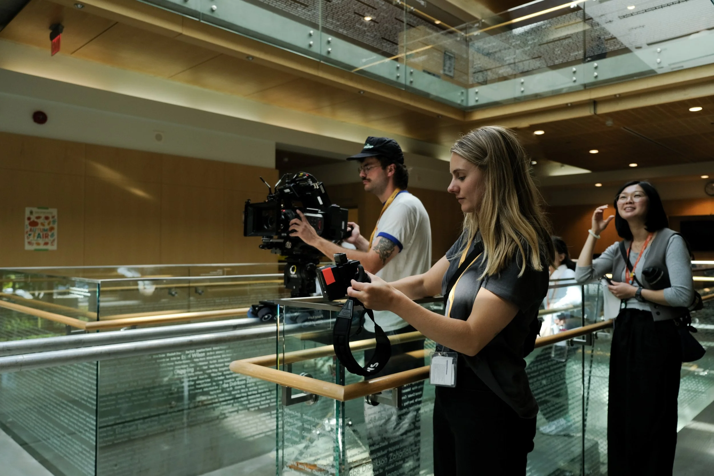 Three people, two women and one man, working on camera equipment at a rail. The woman in the foreground is operating a camera, while the man is adjusting a professional video camera, and the woman in the background is smiling and waving.