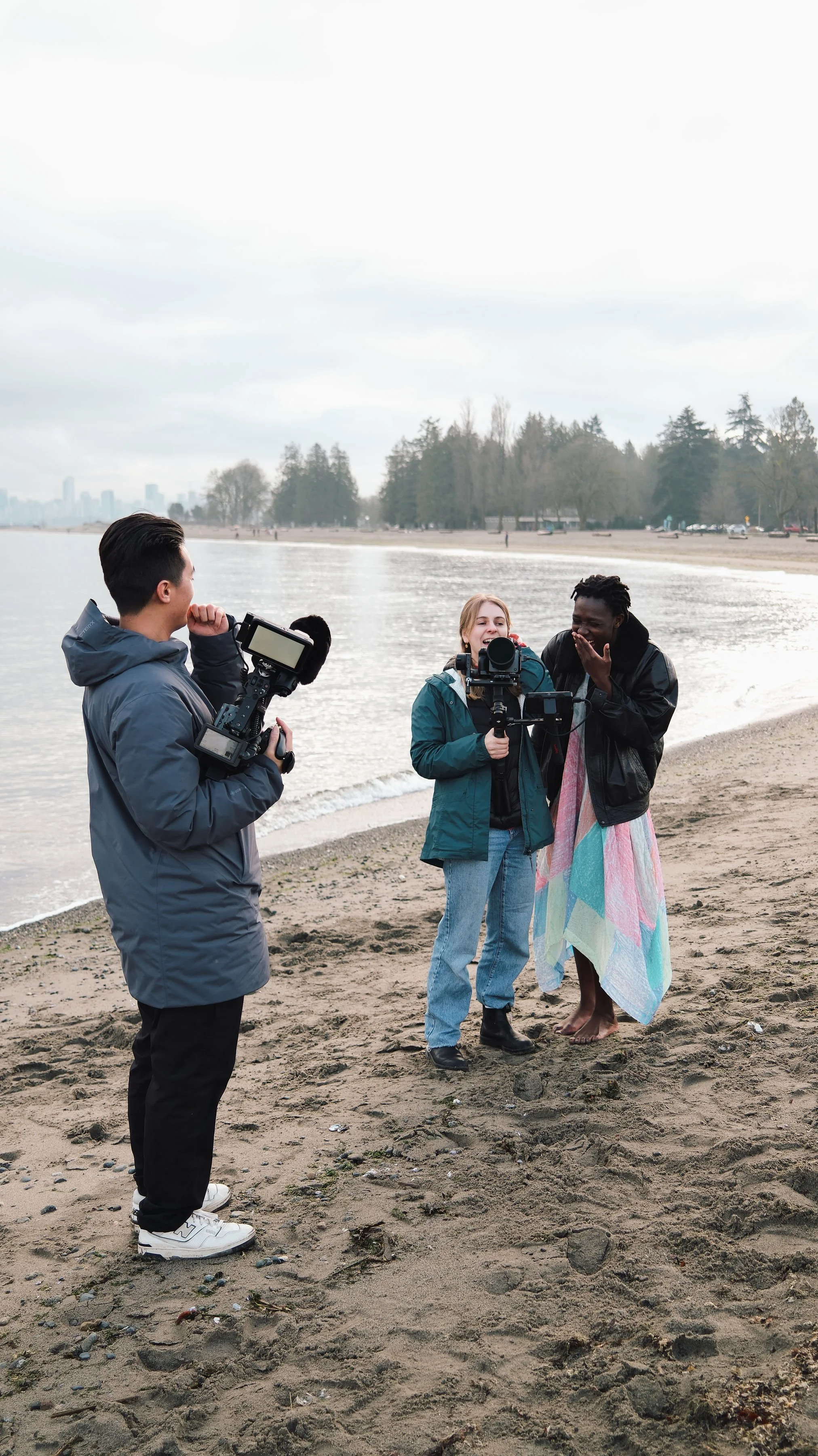 Three people on a beach filming a woman wrapped in a colorful towel, with trees and cityscape in the background.