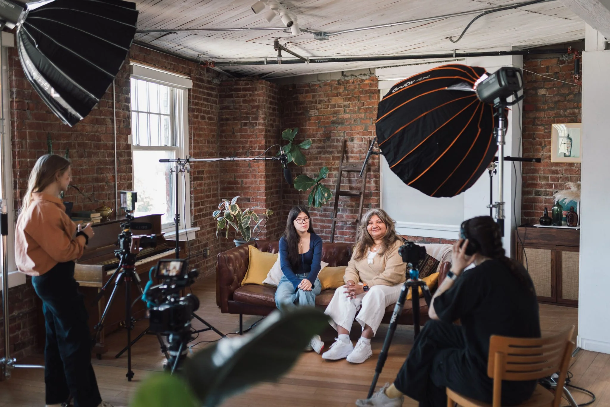 Two women sitting on a brown leather sofa in a brick-walled room, being filmed for an interview or video. Crew members are setting up lights and camera equipment around them, with large studio lights visible.