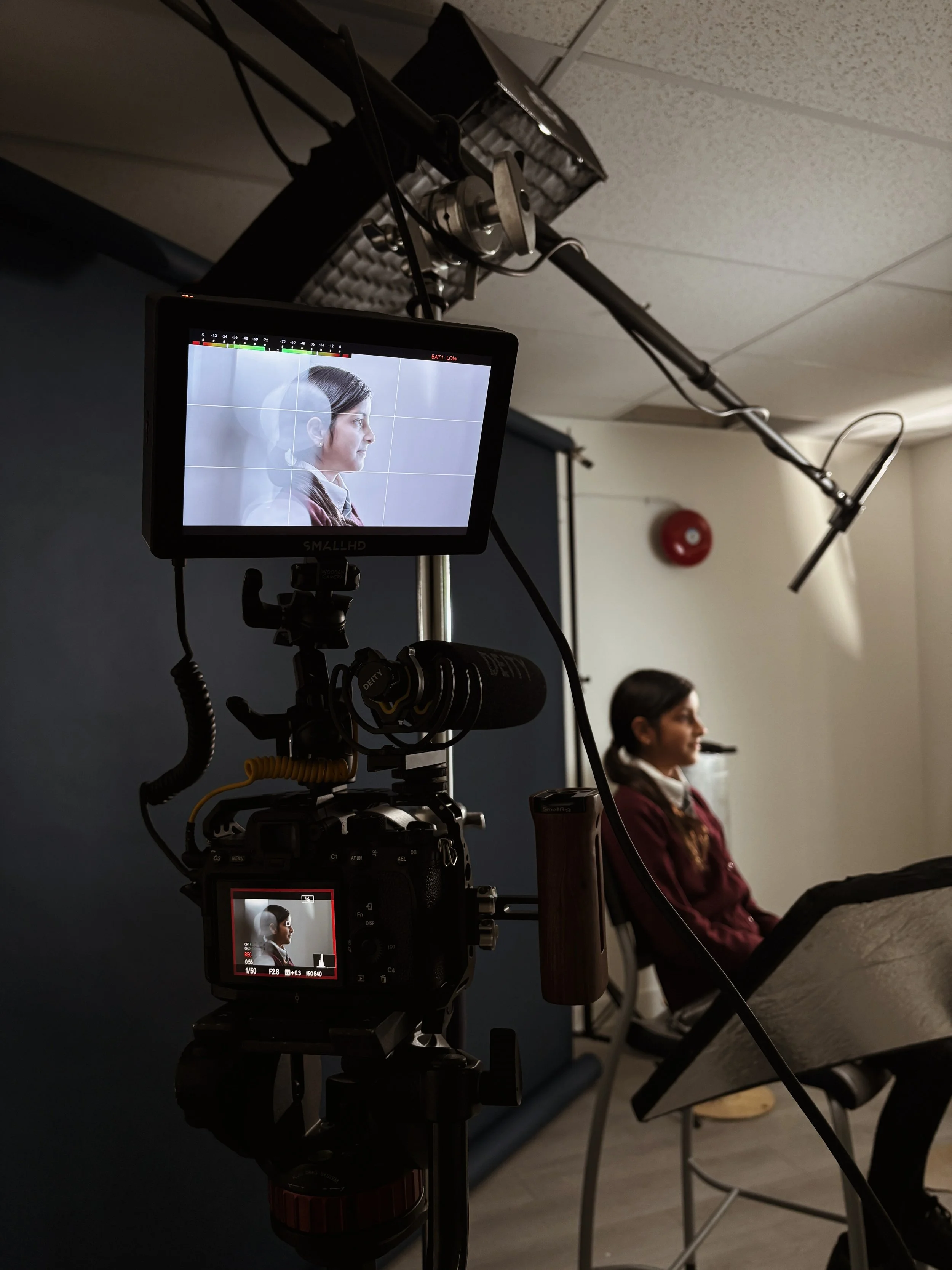 A woman in a maroon sweater sitting in front of a camera on a tripod, with a black backdrop and professional lighting set-up, during a recording or photo shoot.