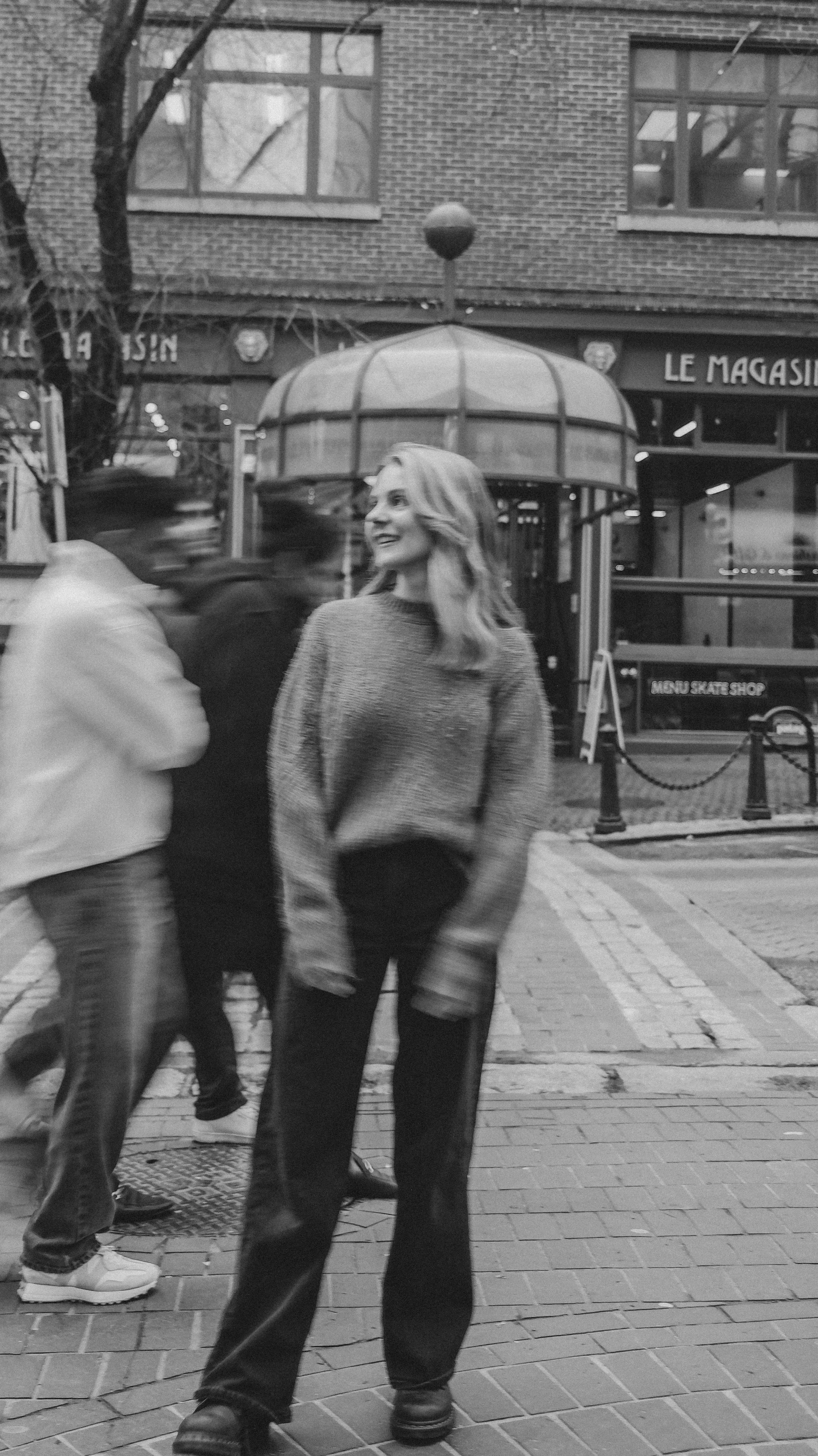 A woman with wavy hair wearing a sweater and pants, standing on a city sidewalk, smiling and looking to her left. There are blurred people walking behind her and buildings with large windows and a sign reading 'LE MAGASIN' in the background.