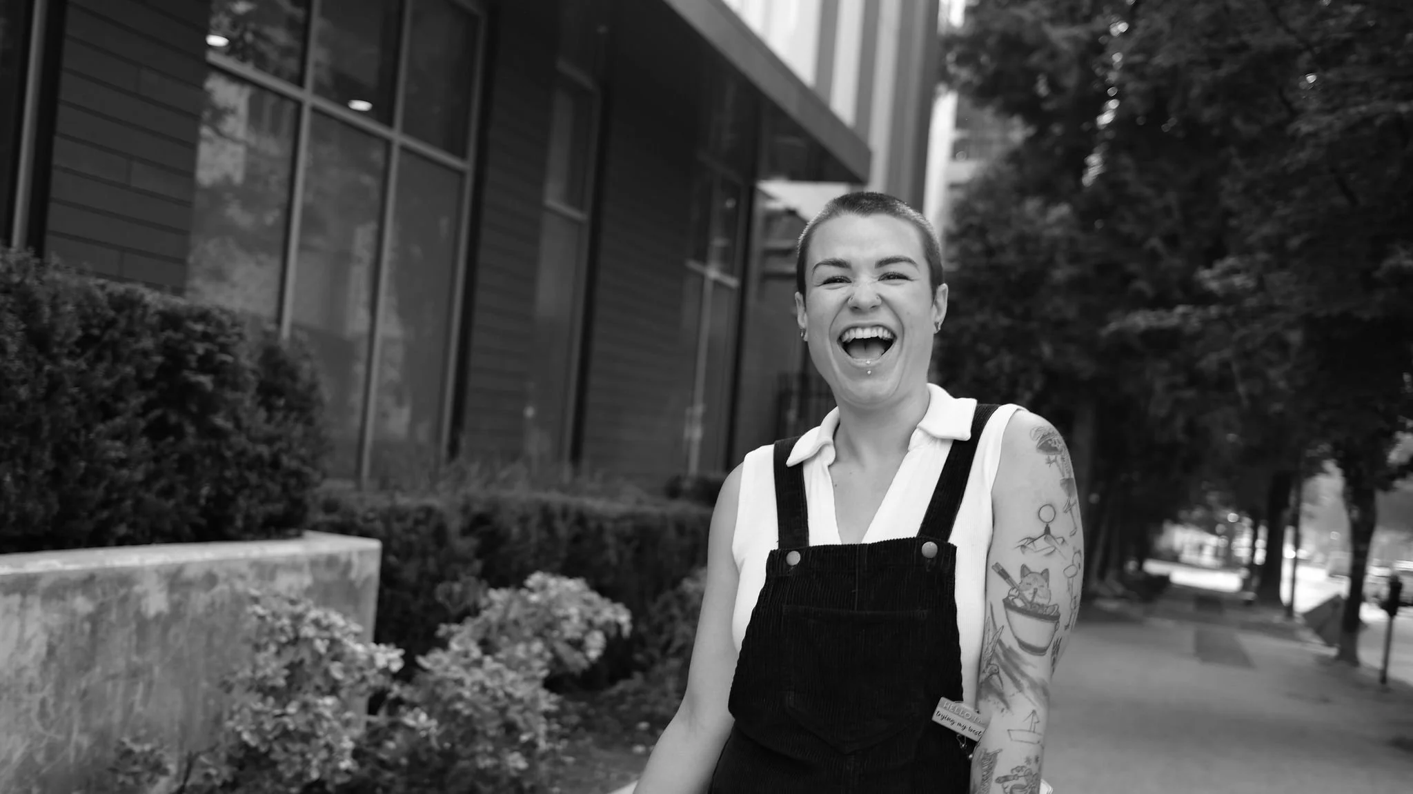 A woman with short hair, tattoos on her arms, and a big smile, standing outdoors in front of a bush and building, laughing.