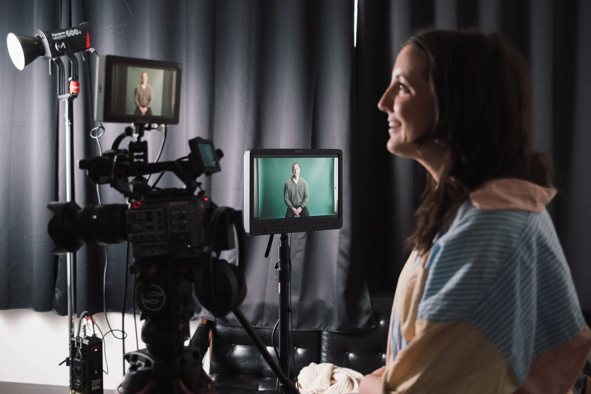 A woman in a colorful sweater is sitting in front of a camera and monitor setup, recording or streaming herself with black curtains in the background.