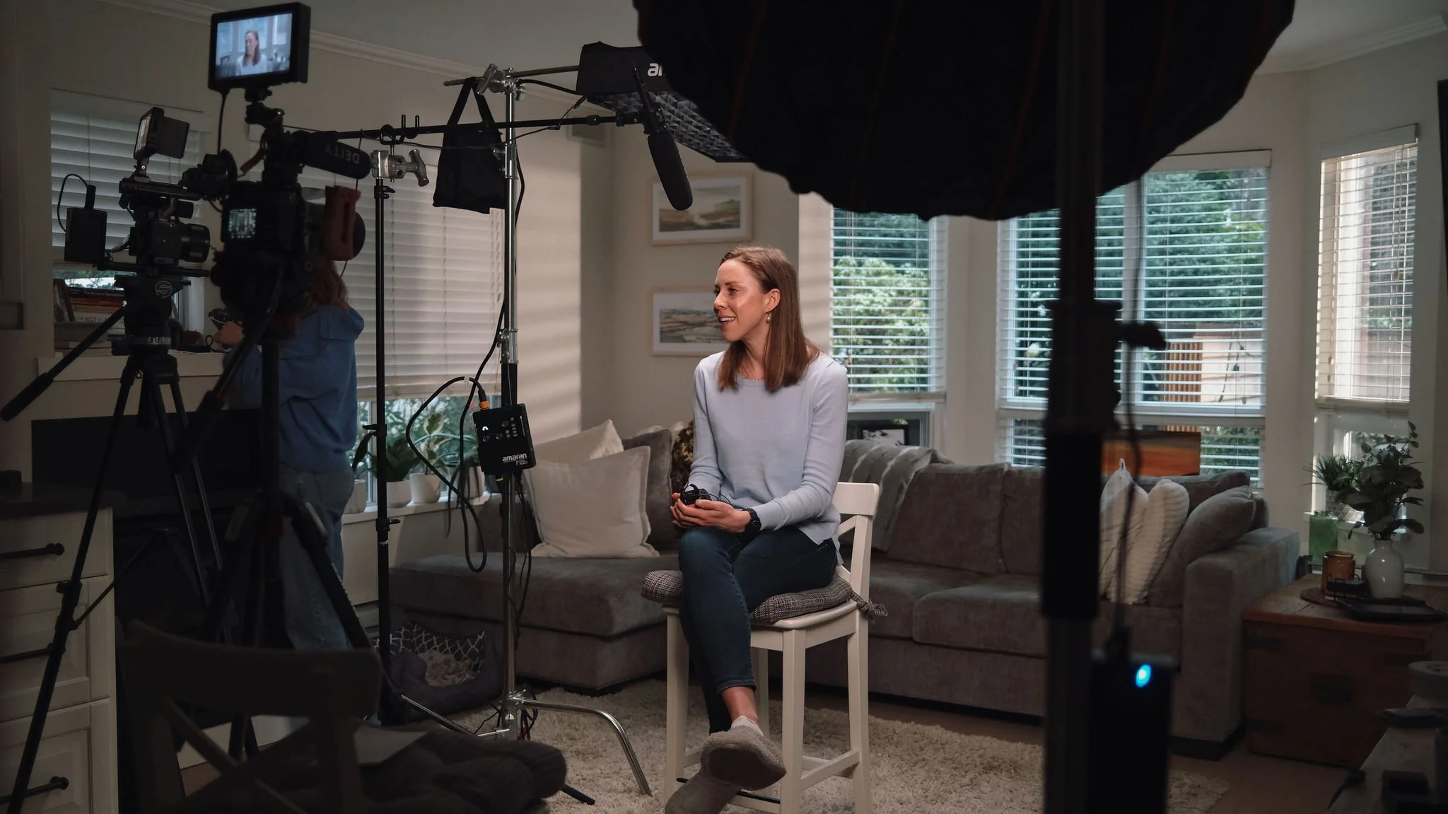 A woman sits on a white chair in a living room, holding a remote control, as she appears to be filming a video or interview. There is professional lighting and camera equipment set up around her, with windows and houseplants in the background.