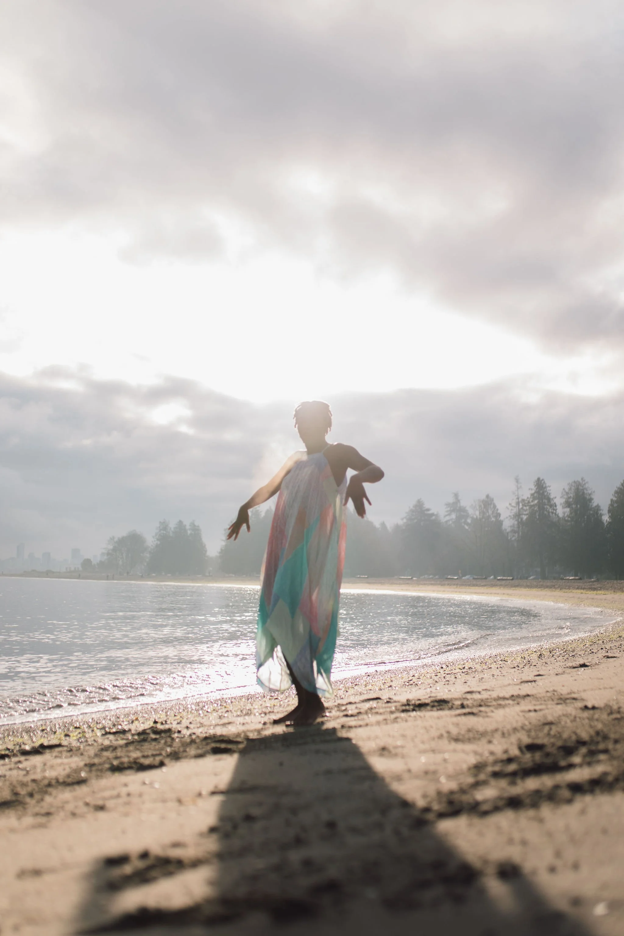 Person walking on the beach during sunset or sunrise, wearing a colorful dress, with the sun partially obscured by clouds in the background.