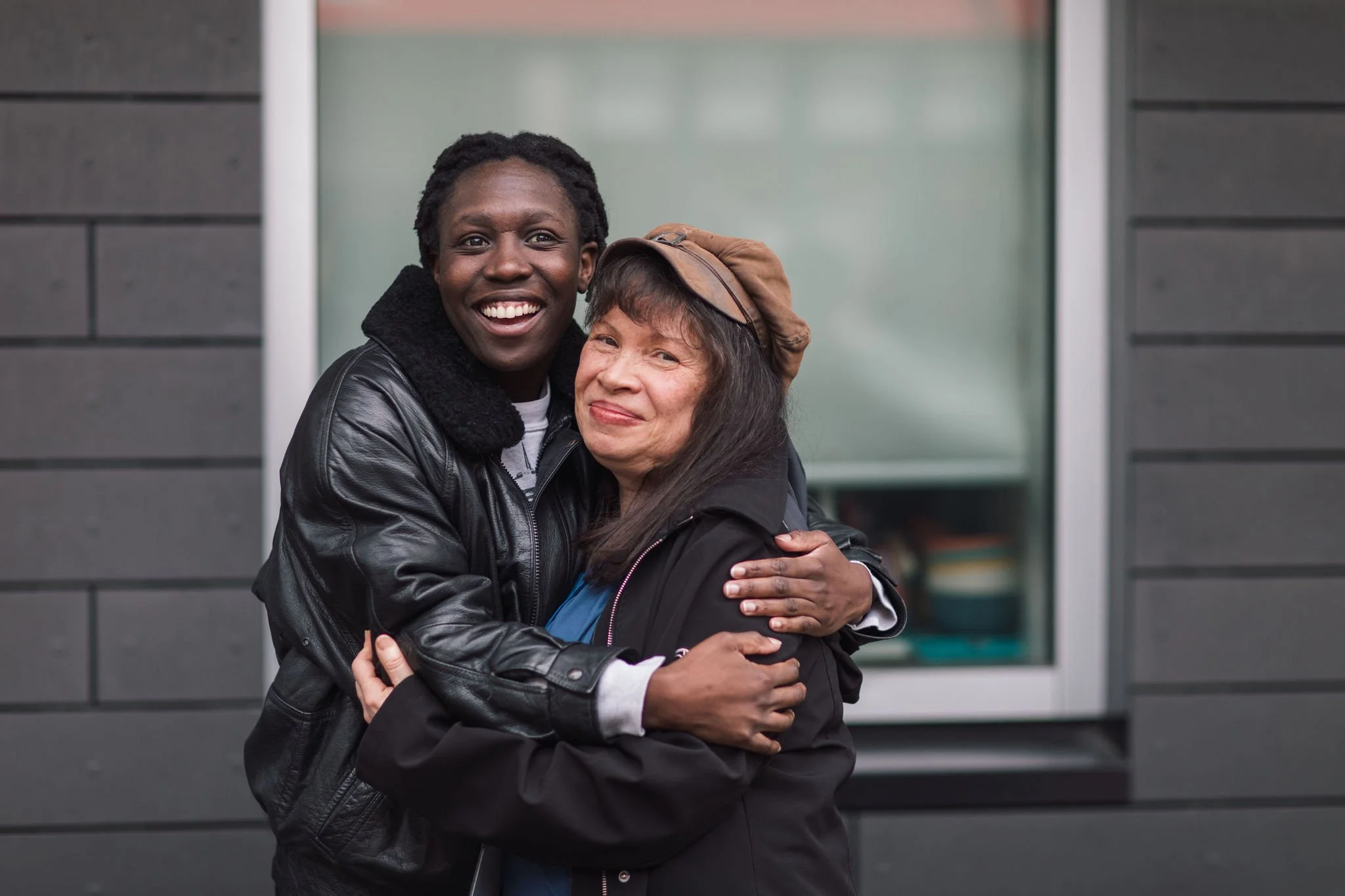 Two women hugging and smiling outdoors near a gray building, one with dark skin and short, curly hair wearing a black leather jacket, the other with lighter skin, long dark hair, and a brown hat, smiling and wearing a black jacket.