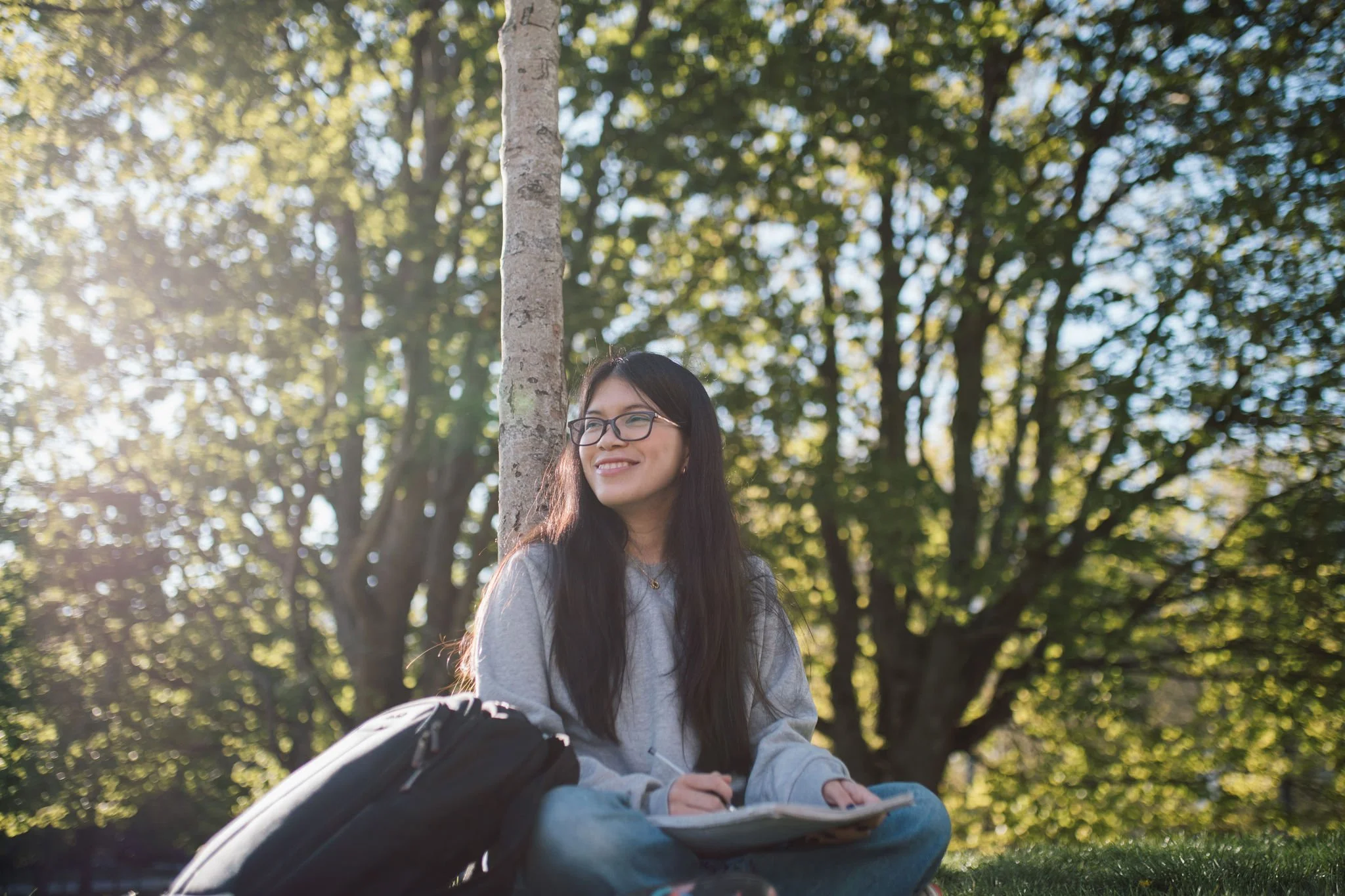 A young woman with glasses, long dark hair, and a gray hoodie sitting outdoors on grass near a tree, smiling and holding a notebook, with a black backpack beside her, during a sunny day with green trees in the background.