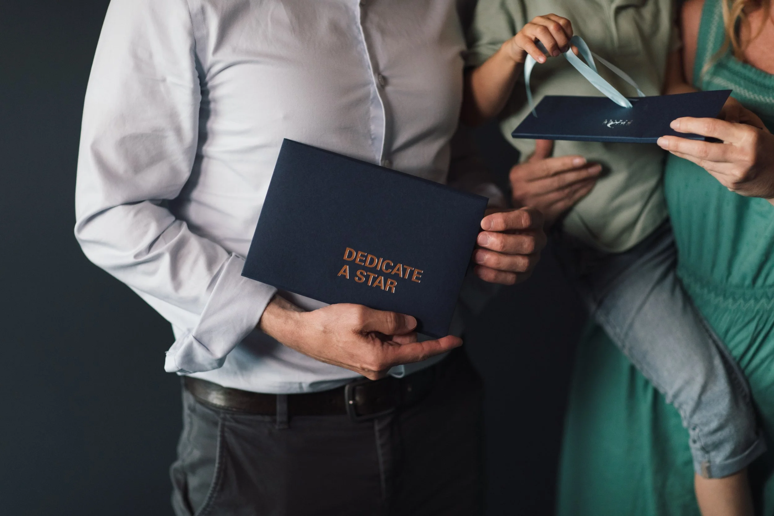 Person holding a navy blue box with the words 'Dedicate A Star' in gold lettering, and people sharing a gift at an event.