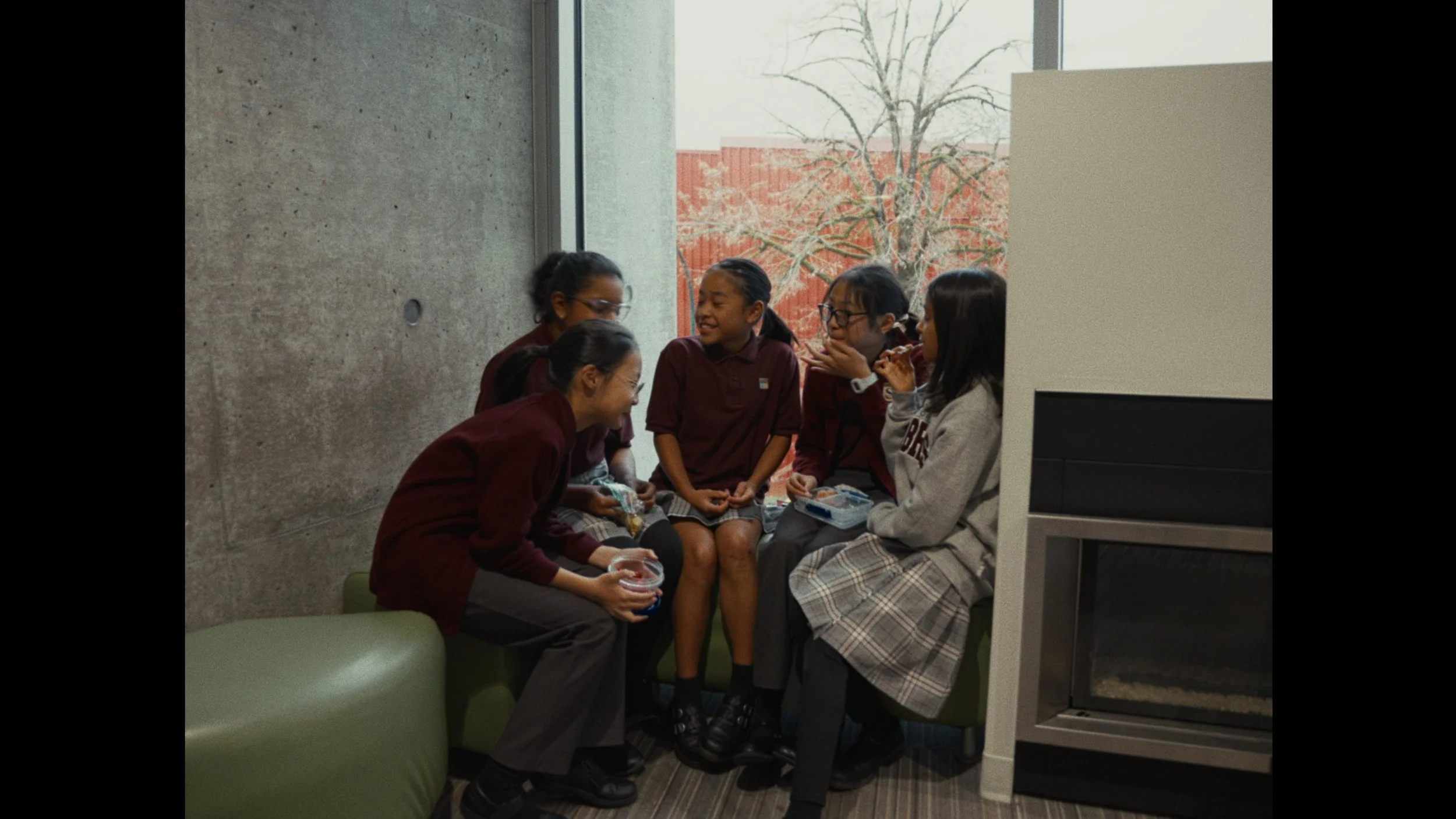 Group of six girls in school uniform sitting on a green couch, laughing and talking. They are in a modern indoor space with a concrete wall and large window showing trees outside.