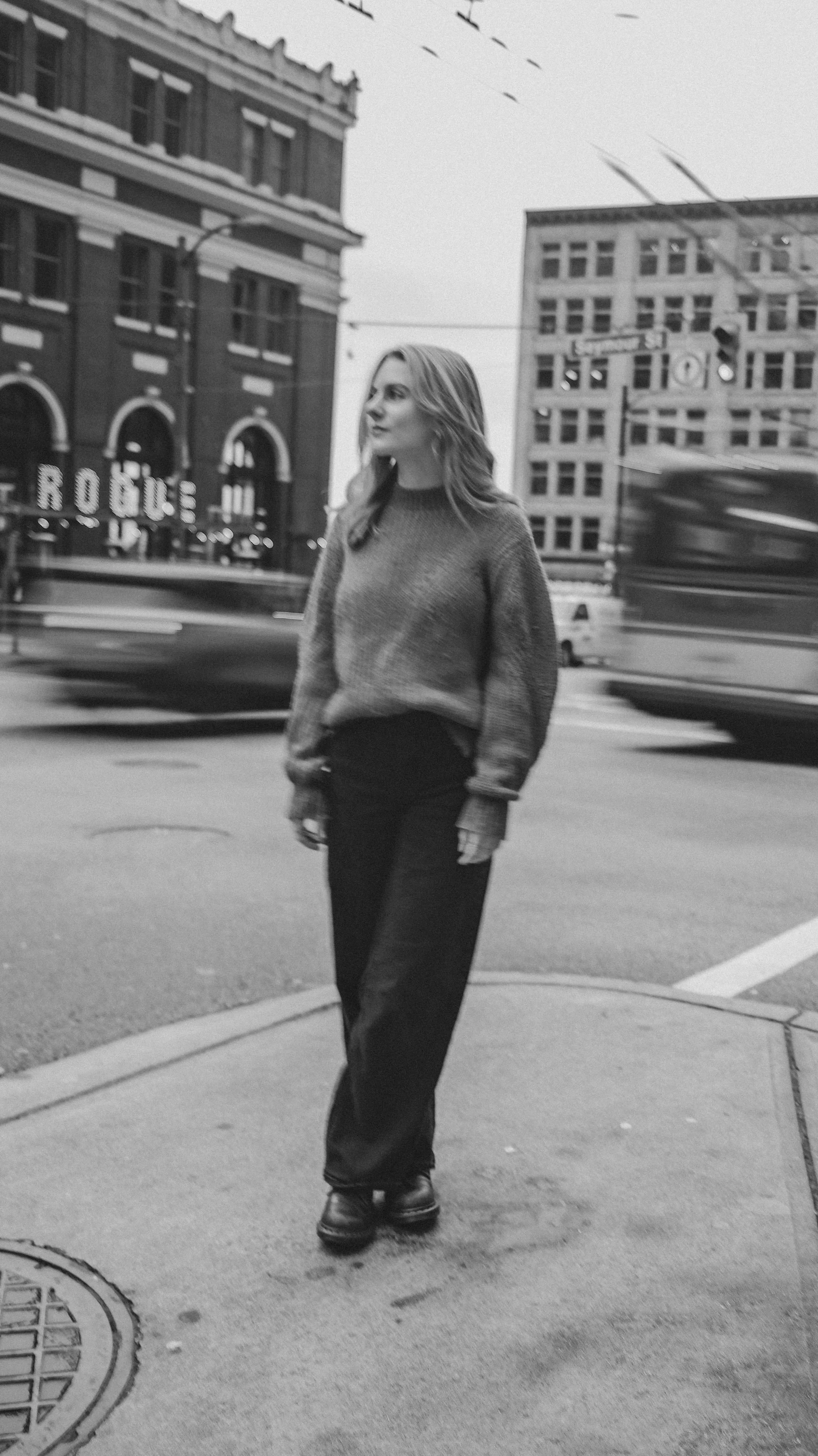 A woman walking on a city sidewalk in black and white, wearing a sweater and wide-leg pants, with blurred city buildings and traffic in the background.
