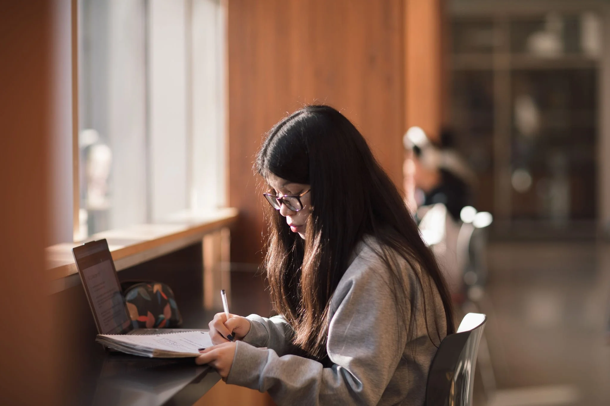 A girl with long dark hair and glasses studying at a wood-paneled library table, writing in a notebook with a pen, illuminated by sunlight coming through large windows.