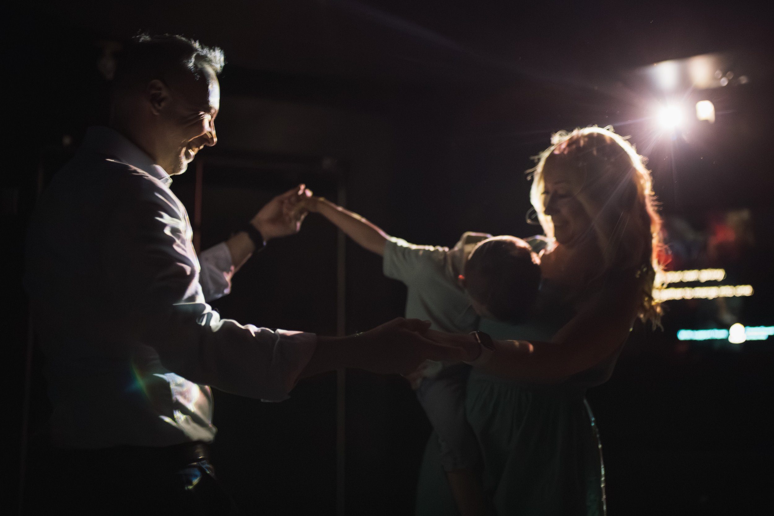 A person holding a baby and dancing with another person, all smiling, backlit by a bright light in a dark setting.