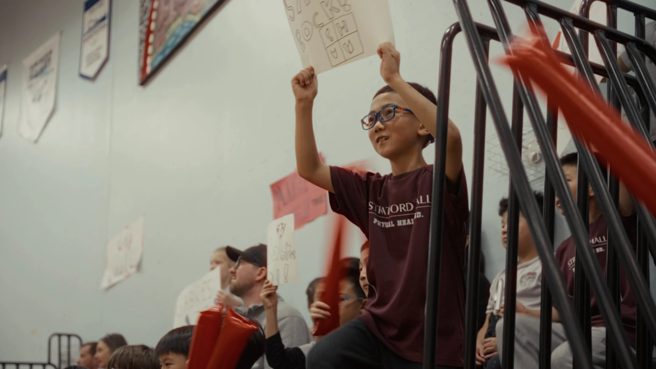 A young boy with glasses holding a sign above his head in a school gym filled with students, some holding signs, as part of a school assembly or event.