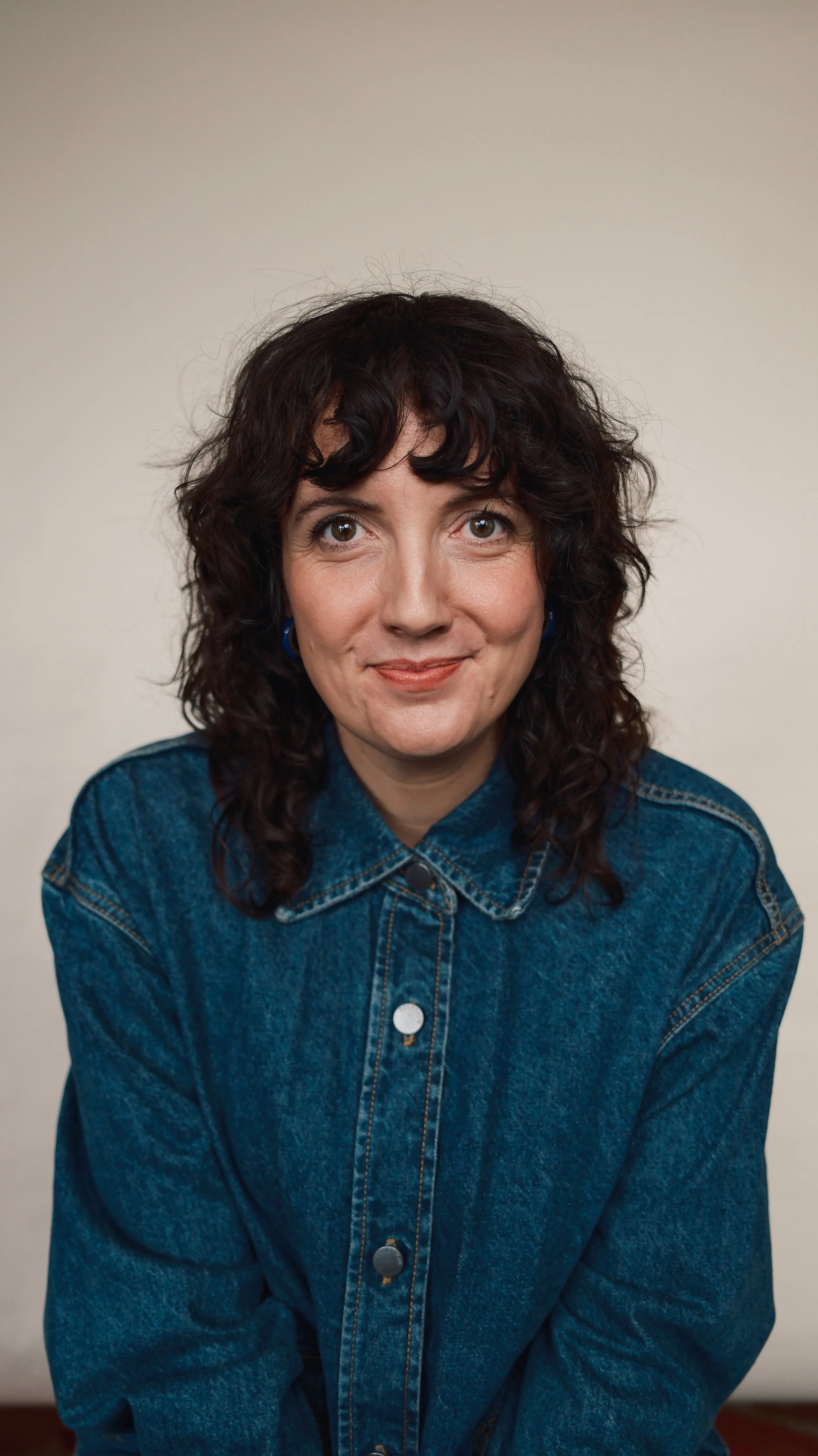 A woman with curly dark hair wearing a denim jacket and blue earrings, smiling softly, against a plain off-white background.