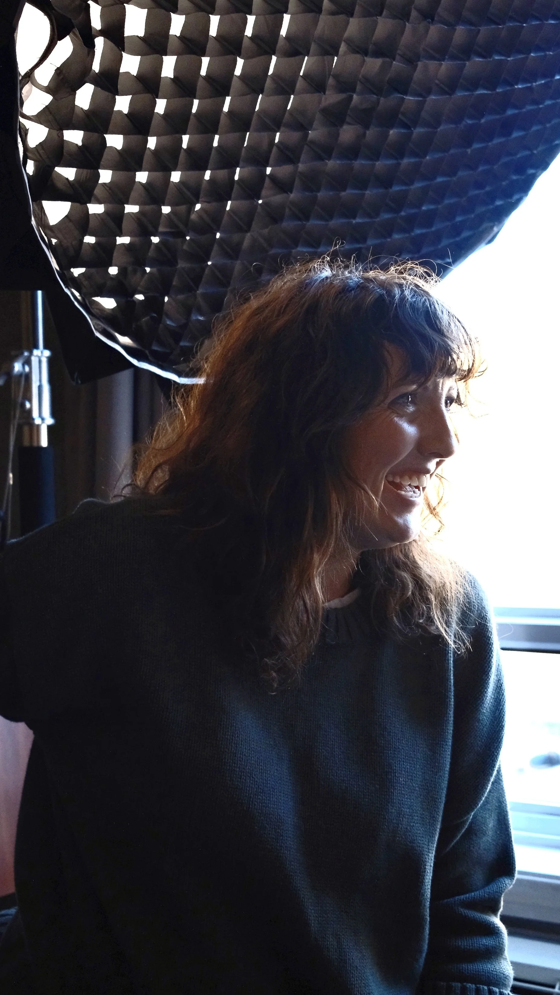 A woman with brown, curly hair smiling during a photography session, seated near a window, with professional lighting equipment angled above her.