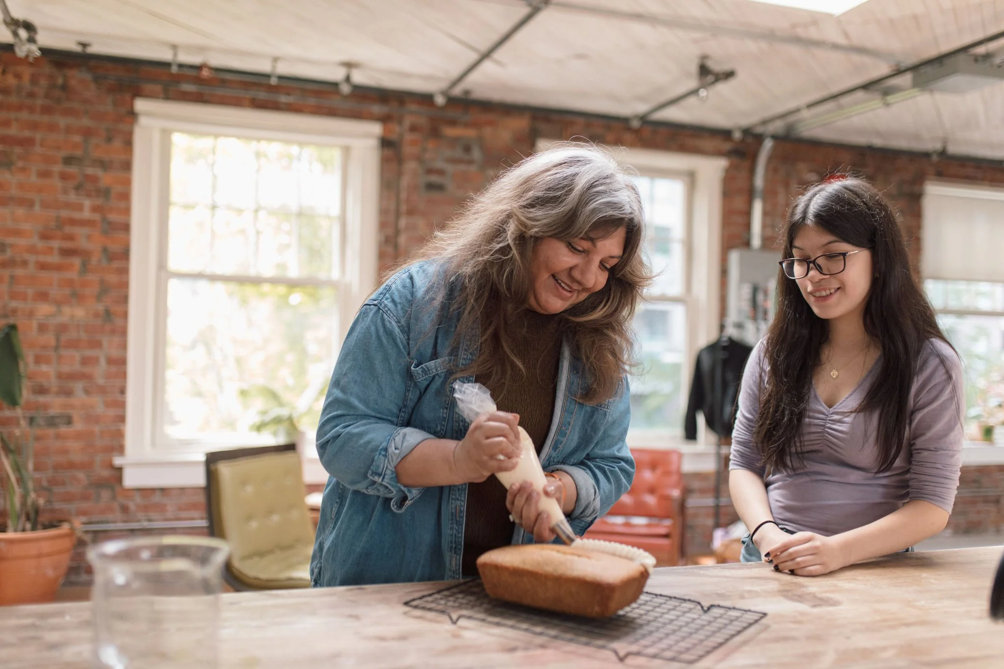 Two women smiling and decorating a loaf of bread in a cozy kitchen with brick walls and large windows.