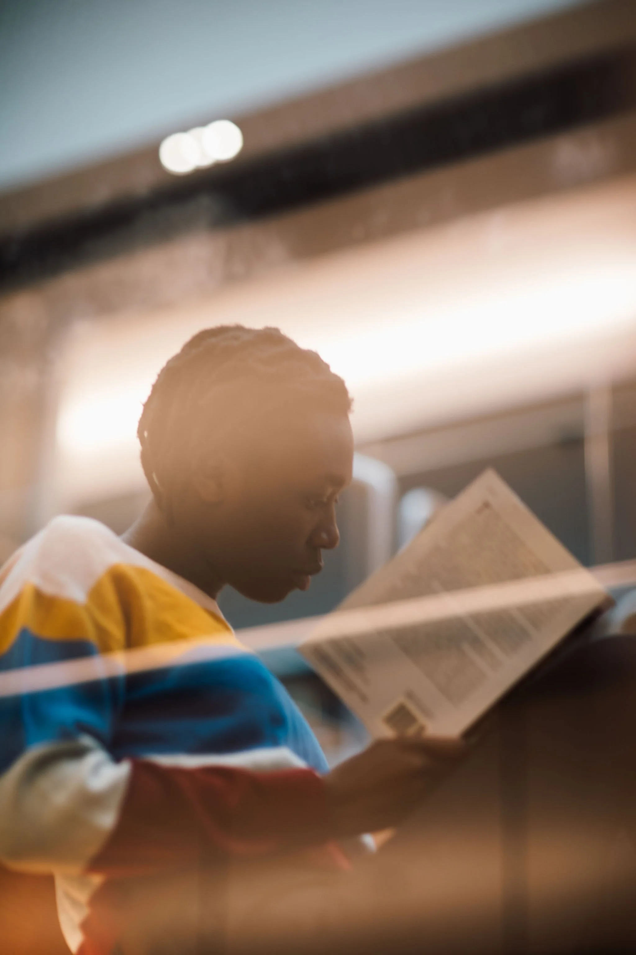 A person with short dreadlocks reading a book, wearing a colorful striped sweater, in a well-lit indoor space.