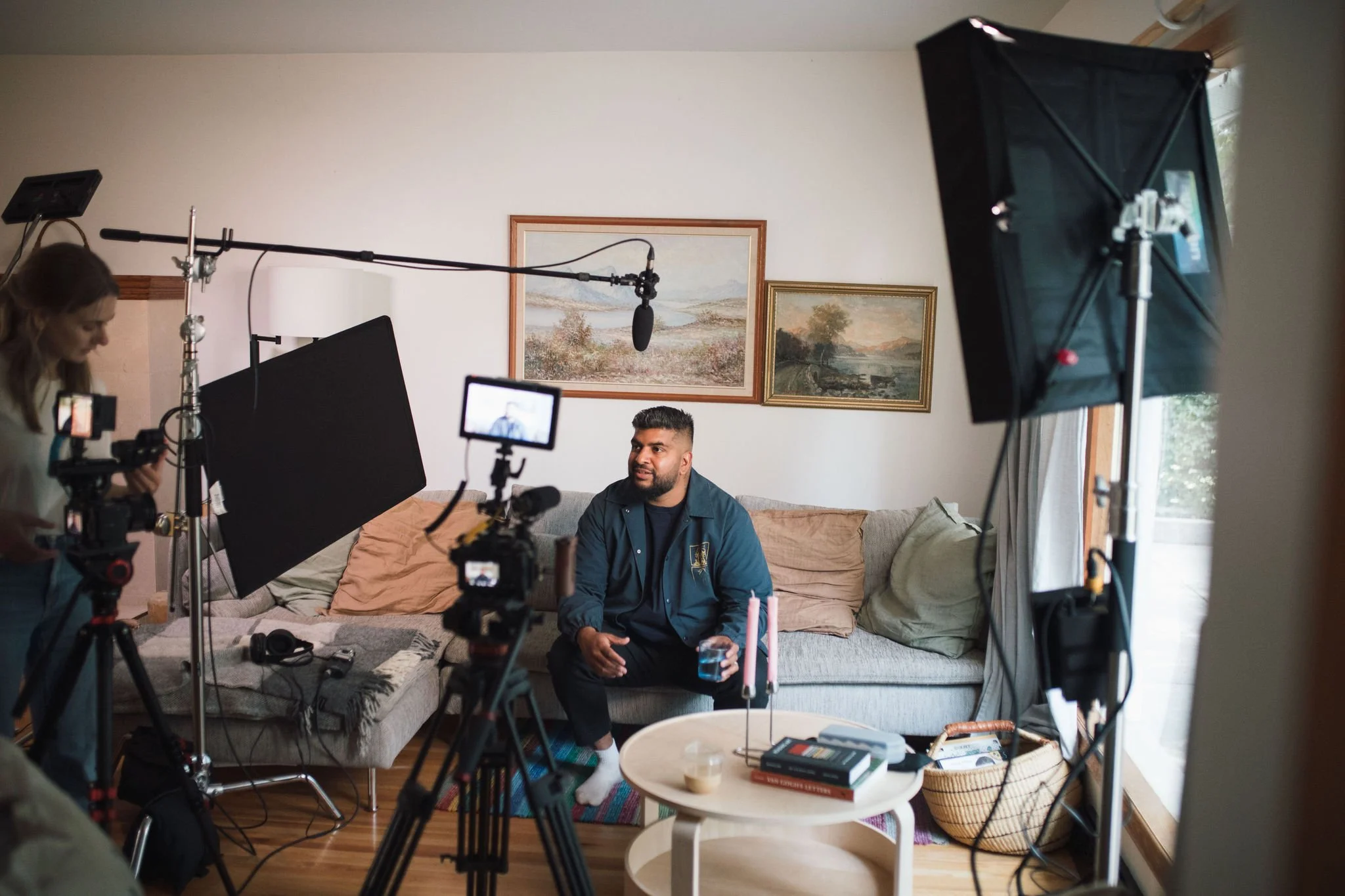 A man with a beard sitting on a couch holding a glass of water during a video recording session. A woman is operating camera equipment on the left. There are lights and microphones set up around him. The room has framed landscape paintings on the wall and a window with curtains.