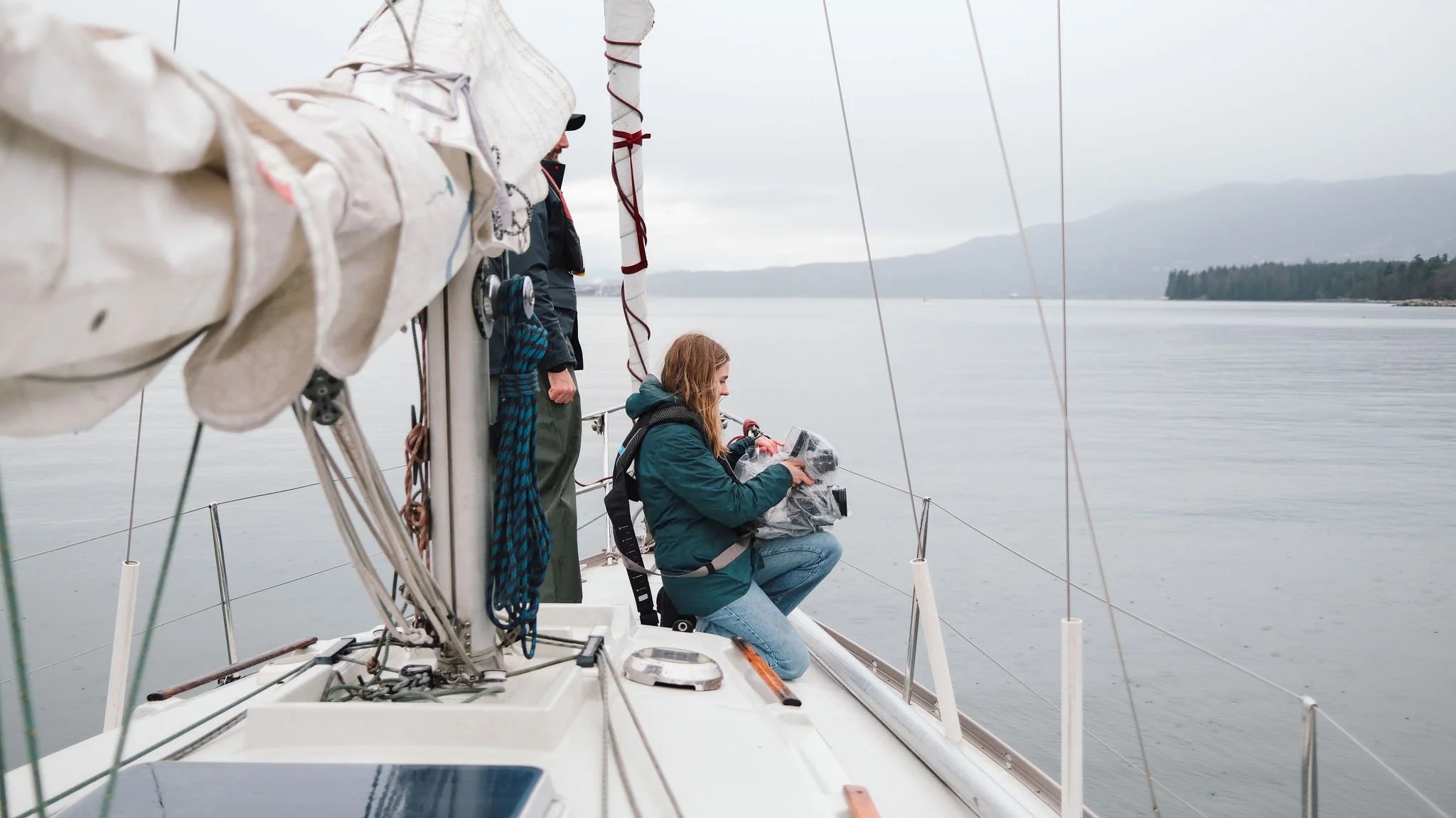 People on a sailboat enjoying a day on the water, with one person sitting and looking at equipment, and others standing near the mast, overlooking a calm body of water and distant land.