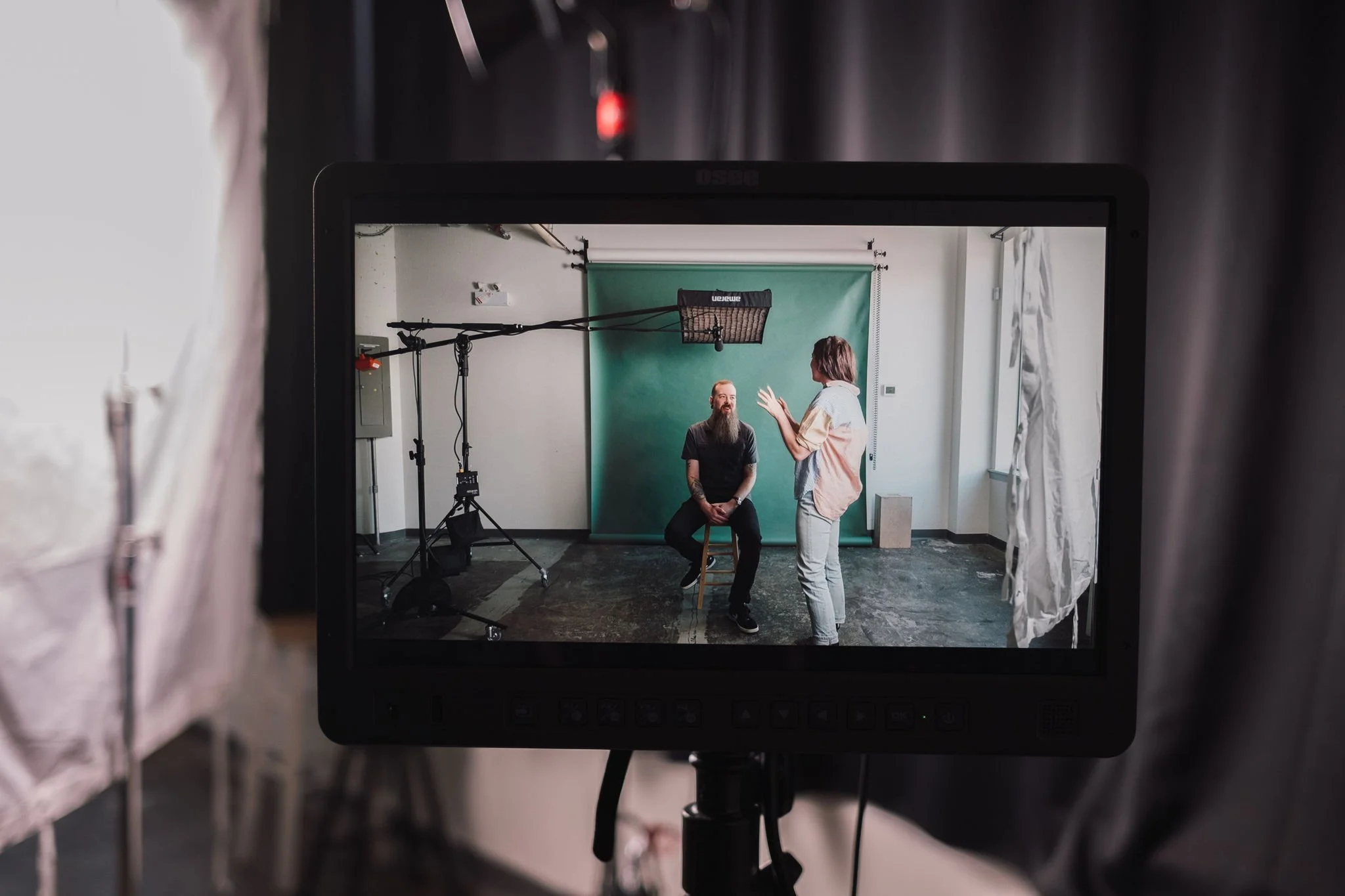 A video camera monitor shows a man with a beard sitting on a stool and talking to a woman standing in front of him in a studio with a green backdrop.