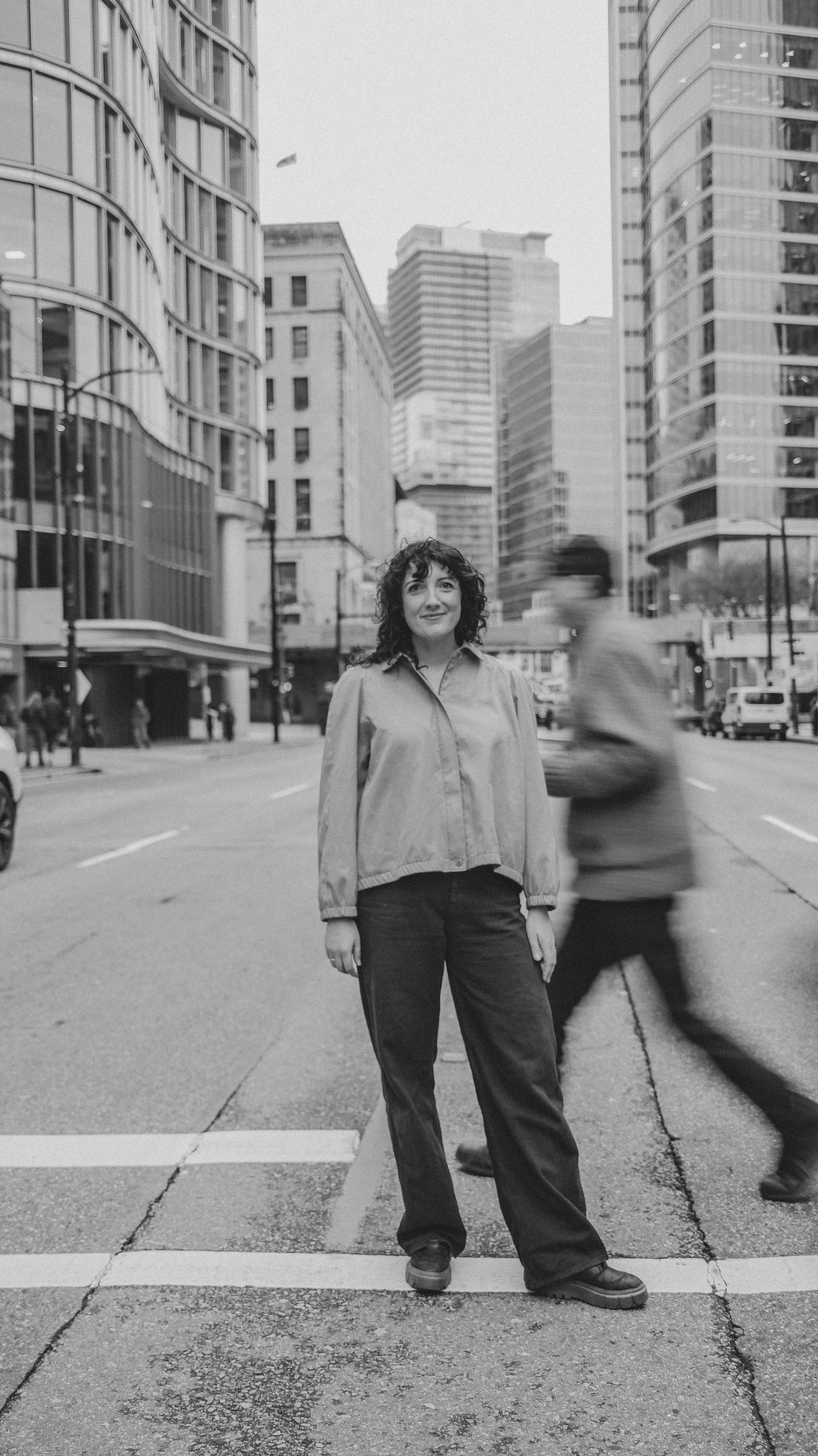 A woman with curly hair, wearing a light jacket and dark pants, standing on a crosswalk in a city street surrounded by tall modern buildings. A blurred person is crossing behind her.