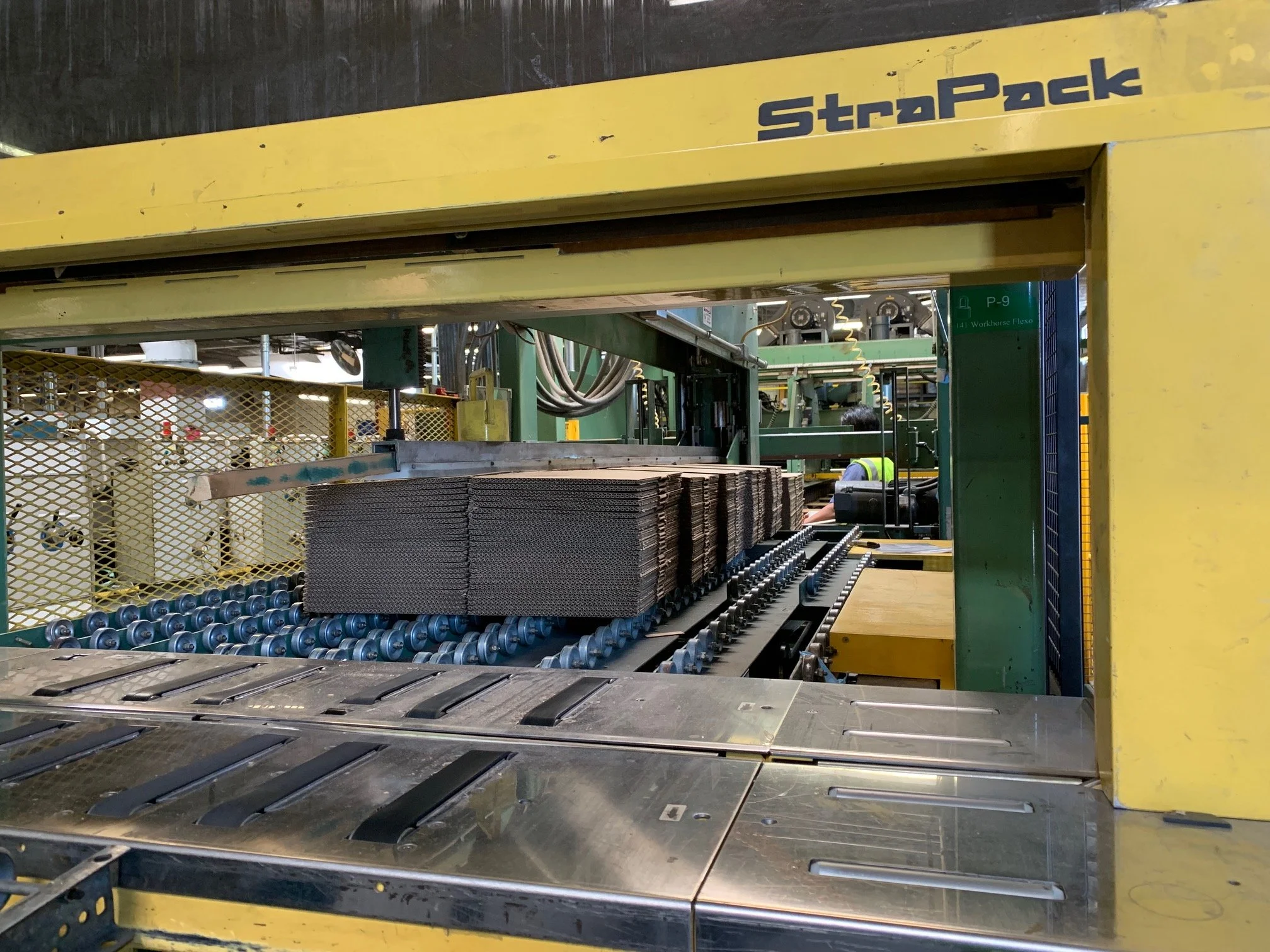Industrial manufacturing machine processing stacked unassembled corrugated boxes with rolling conveyor system.