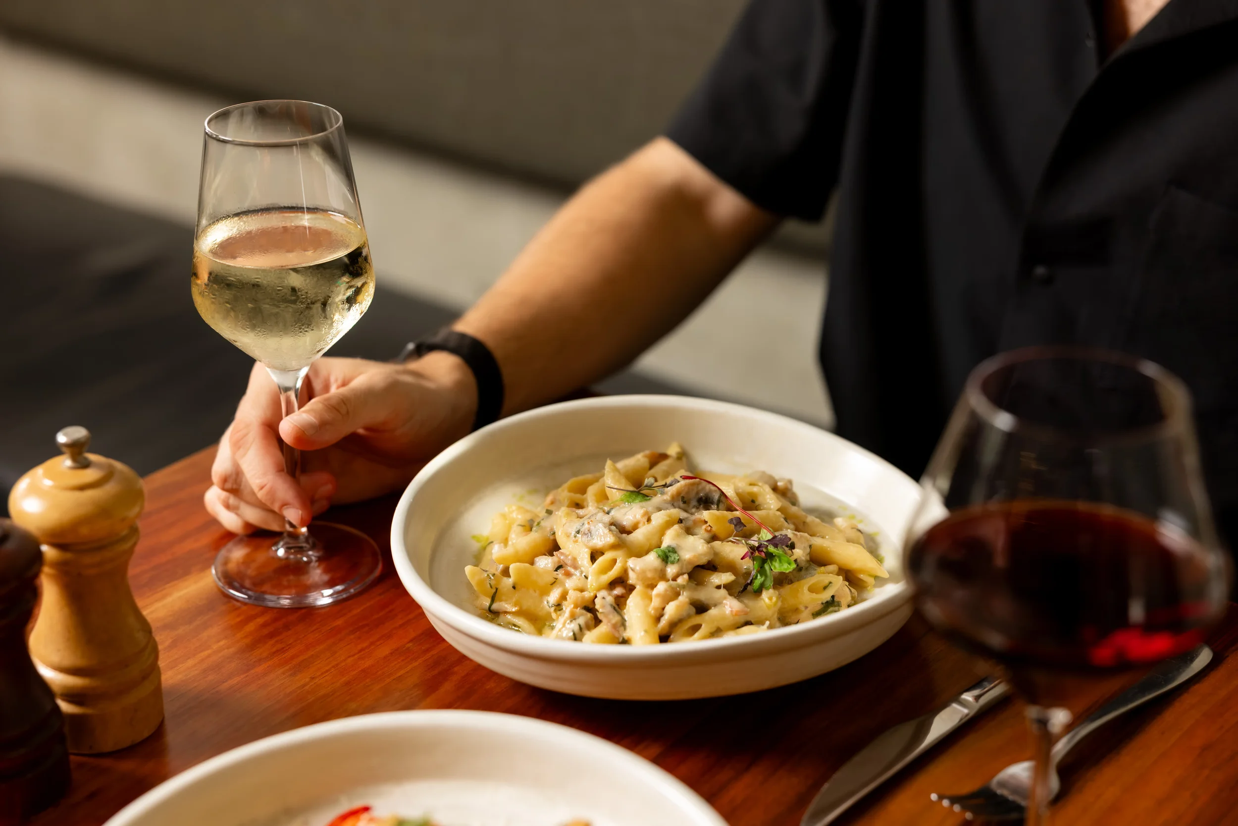 Person holding a glass of white wine with a bowl of pasta on the table, red wine glass, salt and pepper shakers.