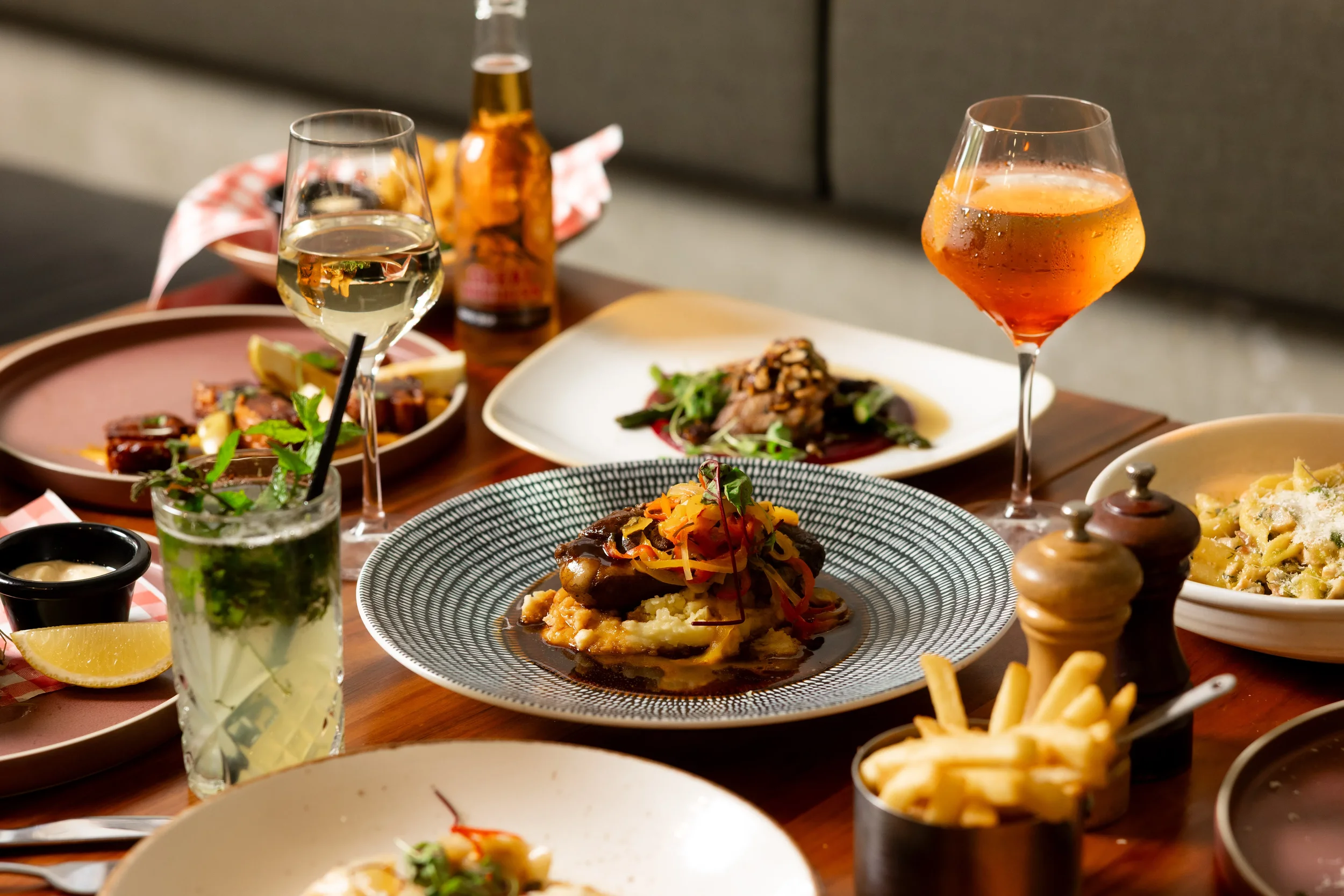A table with various dishes including a beef meal with vegetables, pasta, and two glasses of wine, one white and one red, along with a beer bottle and utensils.