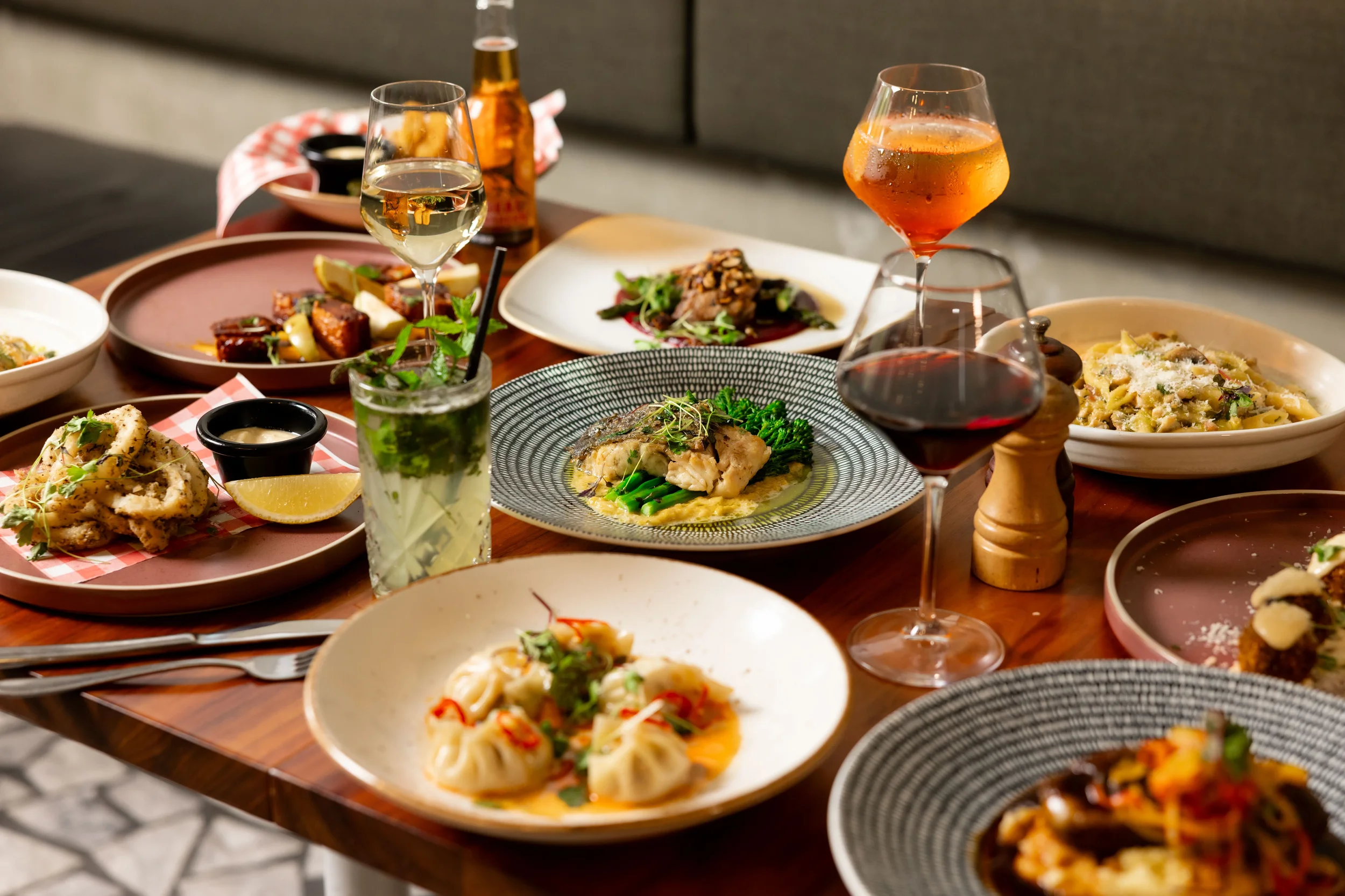 A table set with various plates of gourmet Mediterranean dishes, including pasta, dumplings, seafood, and salads, along with wine glasses, a cocktail, and beer bottles.