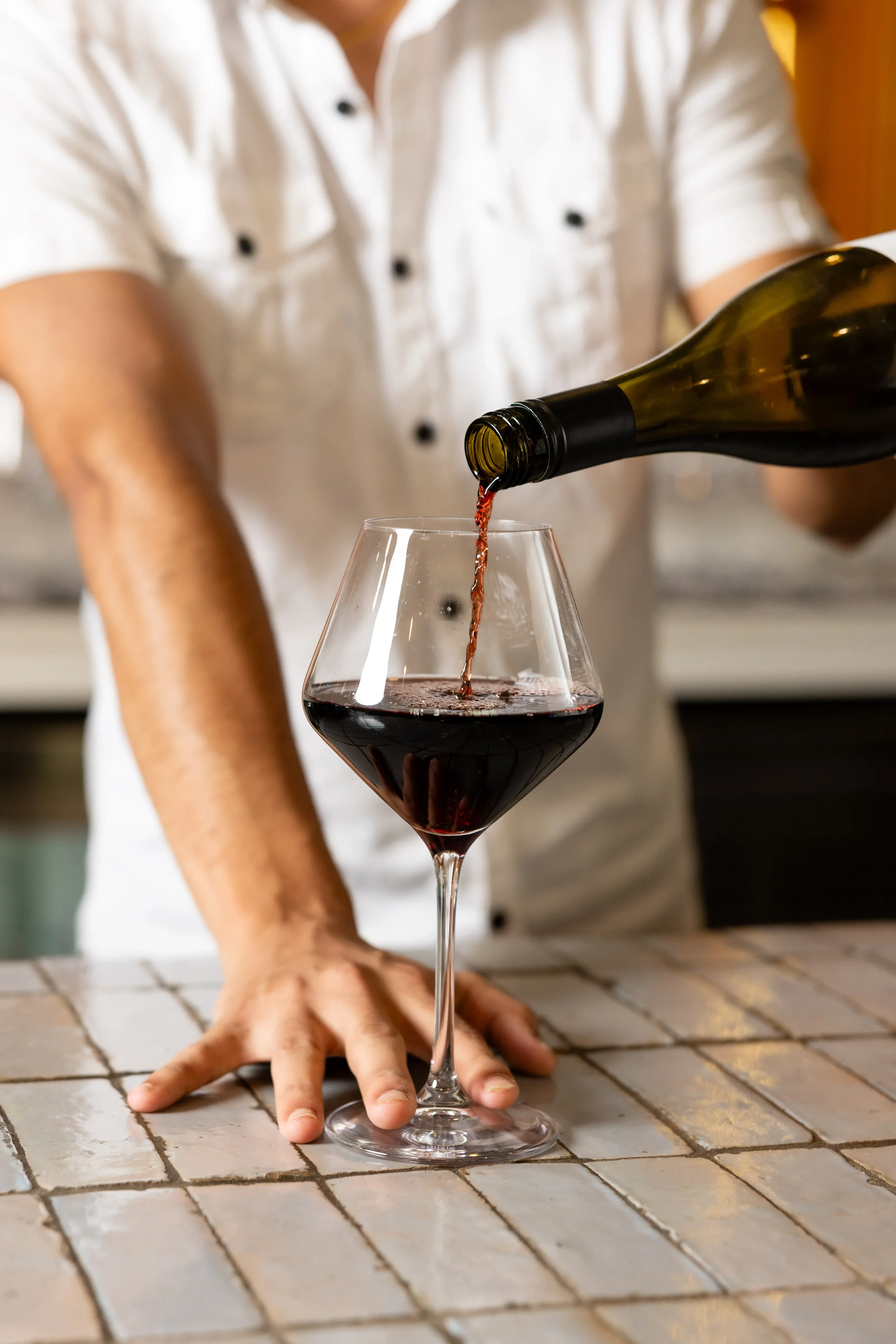 Person pouring red wine into a wine glass on a tiled surface.