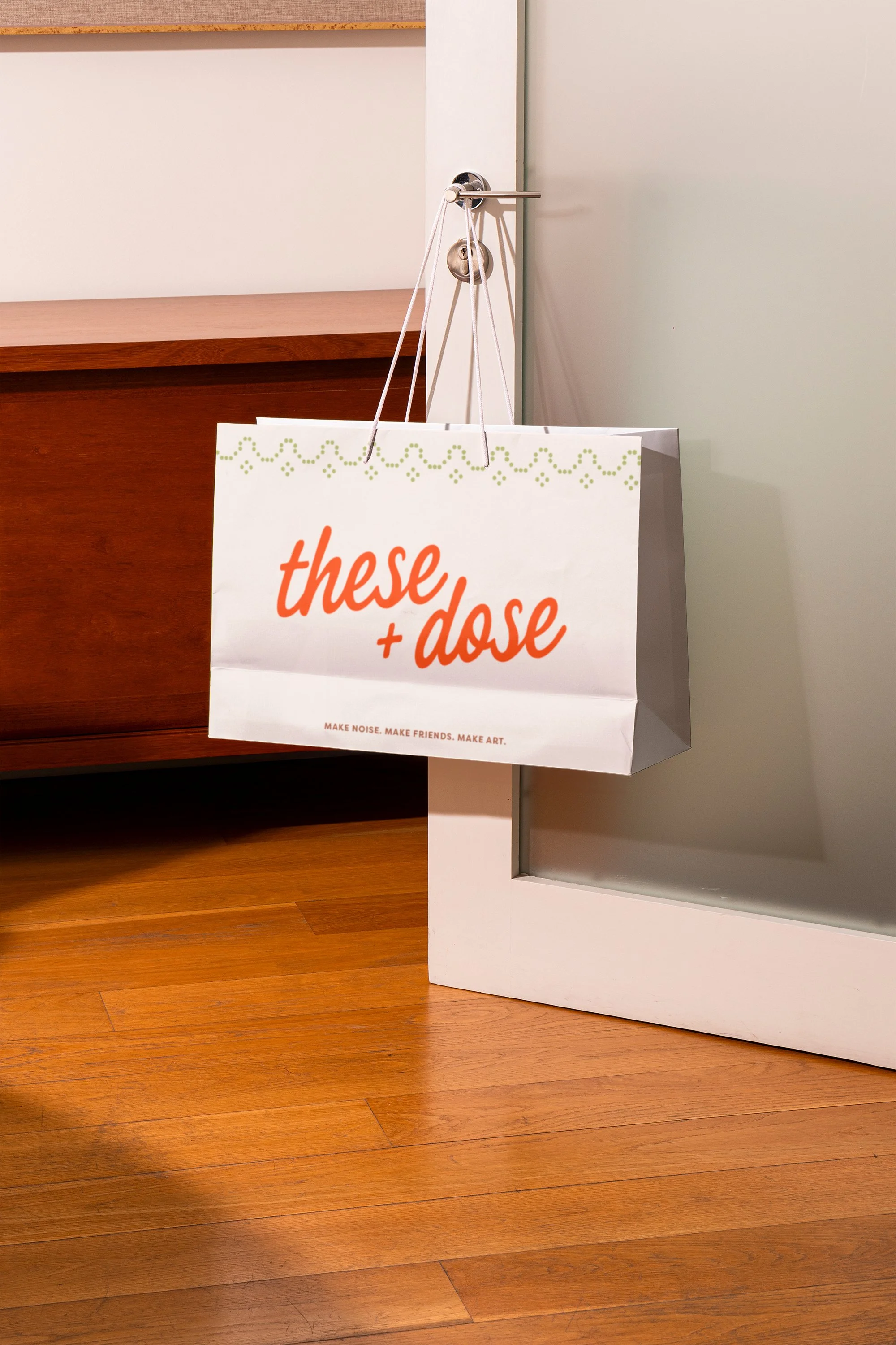 A paper shopping bag hanging on a door handle, with the words 'these + dose' written in orange text and the phrase 'MAKE NOISE. MAKE FRIENDS. MAKE ART.' printed below.