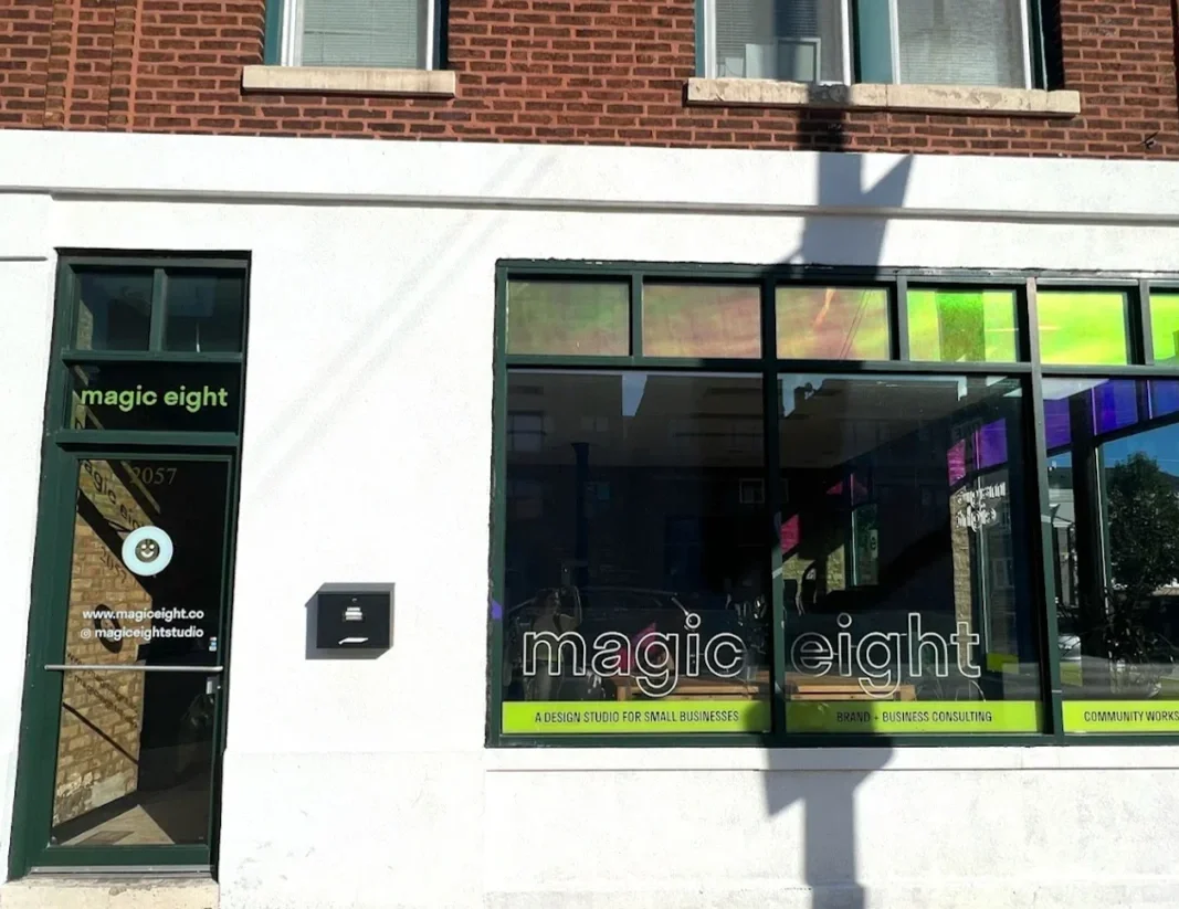 Magic Eight Co. storefront with green and black sign and a window with the same logo, website, and social media handle, along with signs indicating services for small businesses, branding, business consulting, and community work.