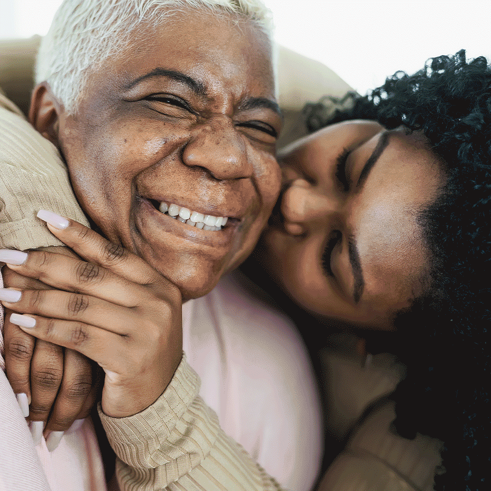 An elderly woman and a young woman share a joyful hug, smiling with their eyes closed.